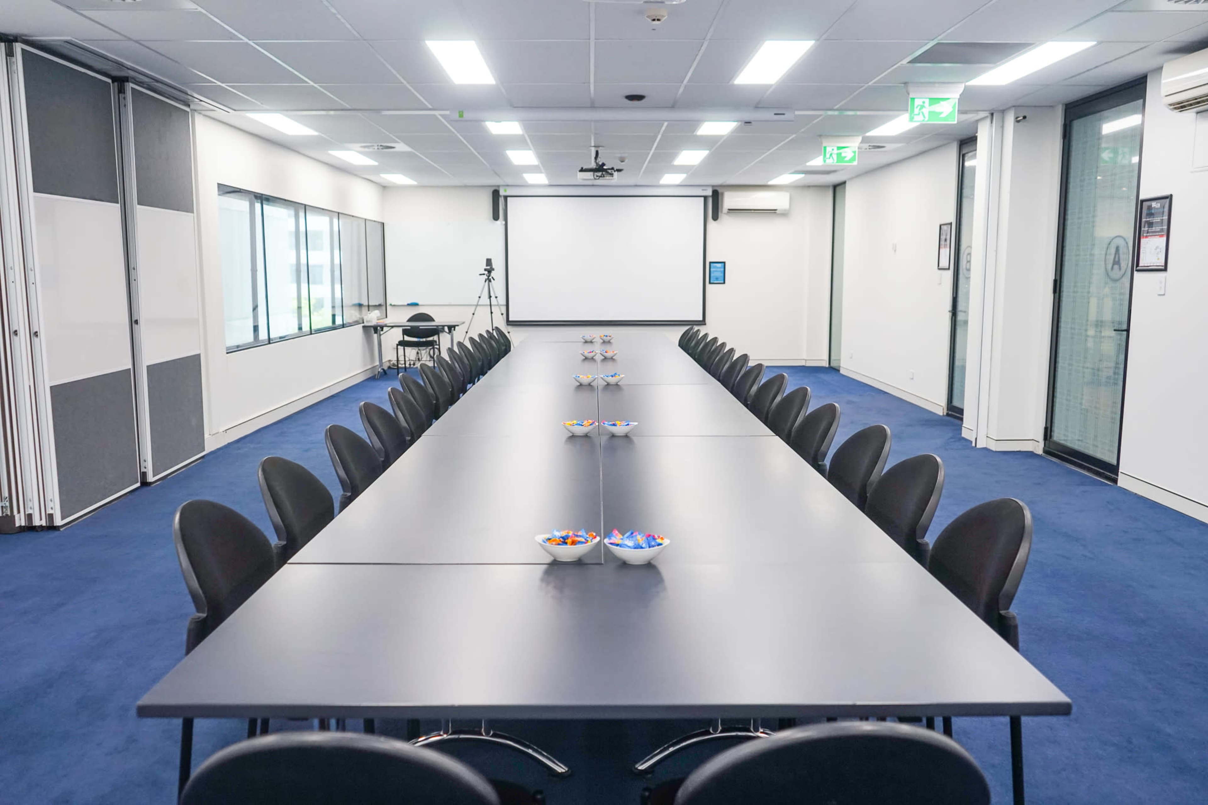 A long conference table with black chairs is set up in a modern meeting room, featuring bowls of snacks at each end and a projector screen at the front.