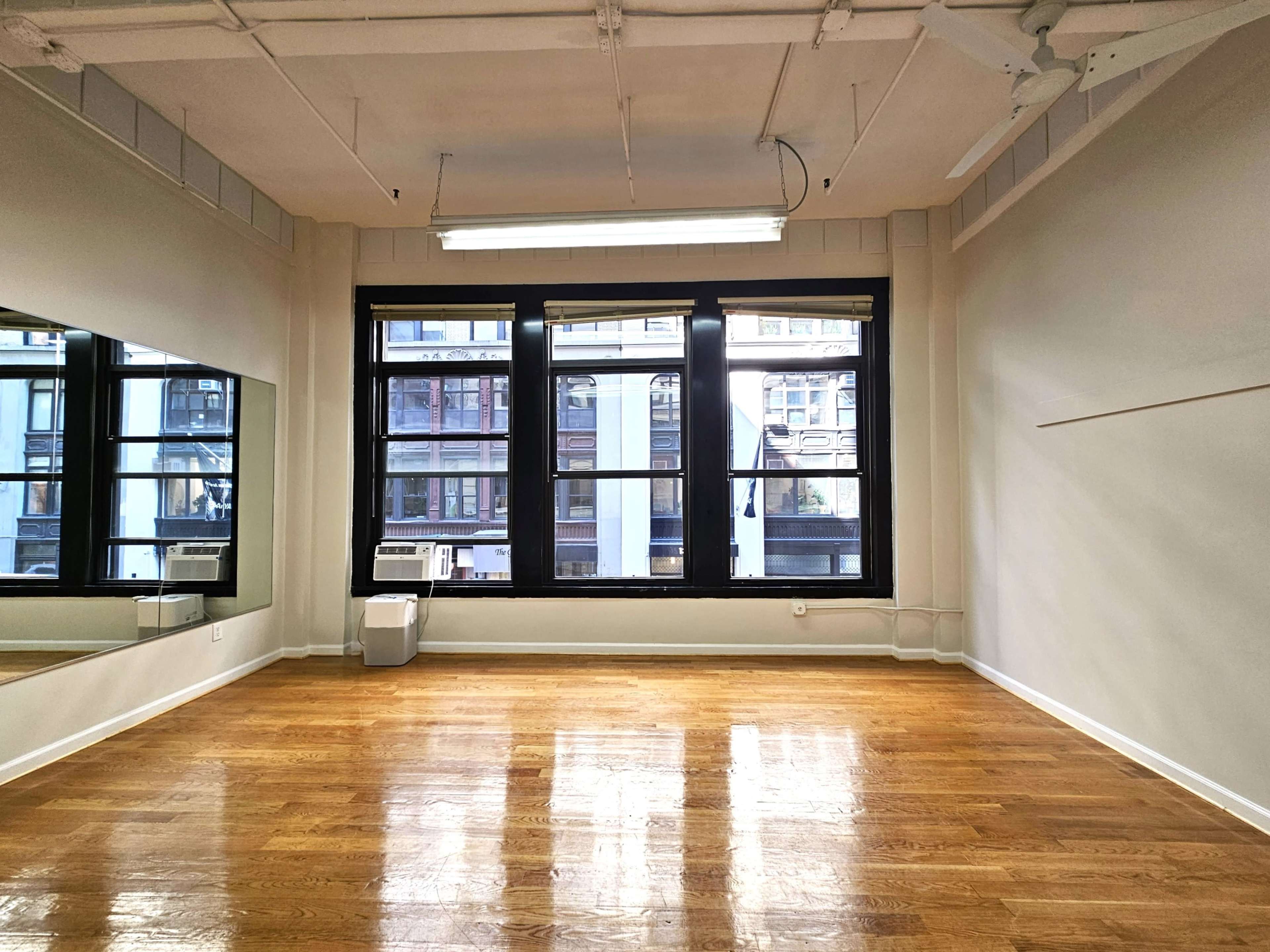 The image shows a spacious room with hardwood floors, large windows, and a wall-mounted air conditioning unit.