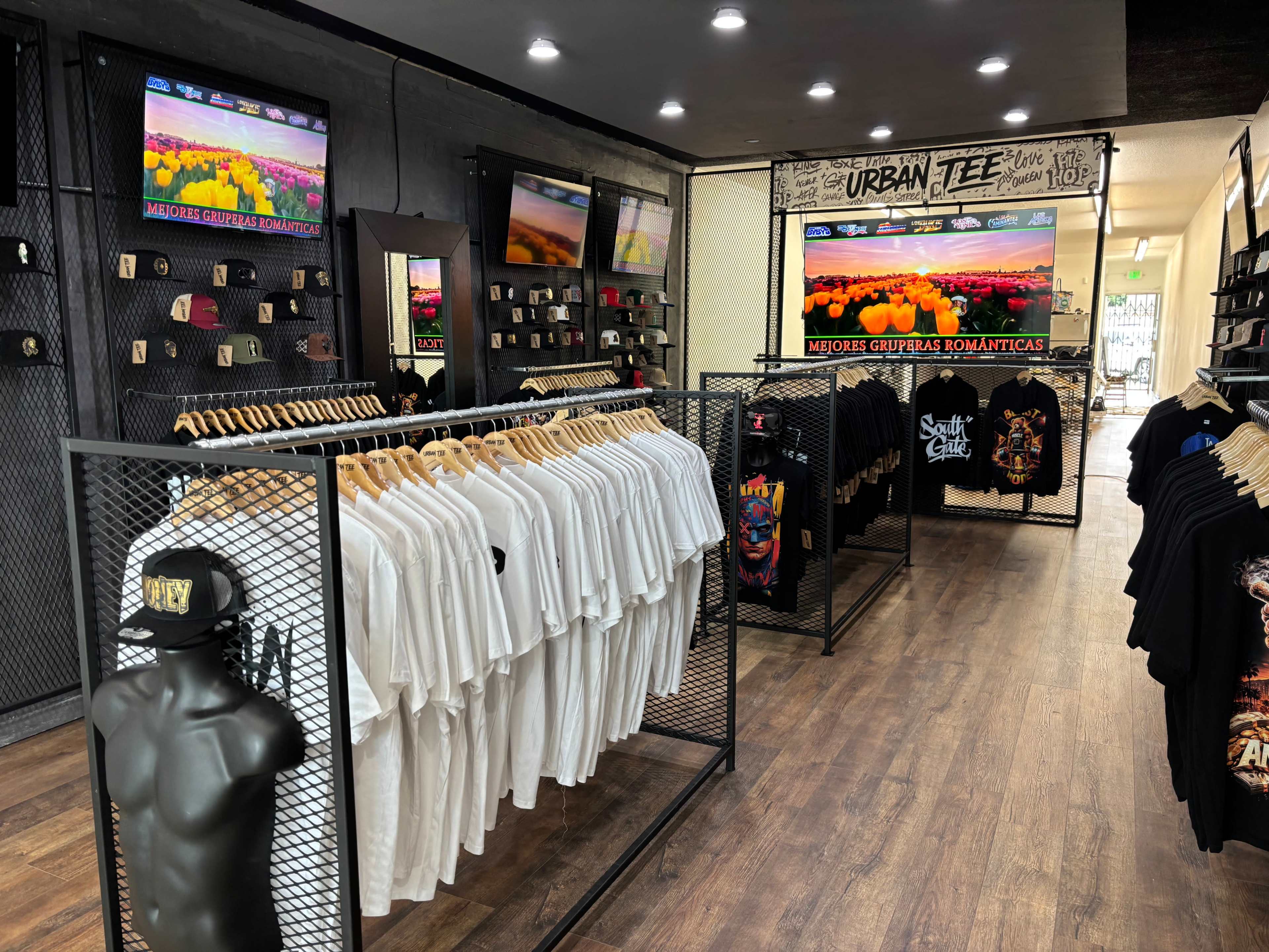 The image shows an apparel store featuring racks of T-shirts and hats arranged neatly, with digital screens displaying colorful graphics in the background.