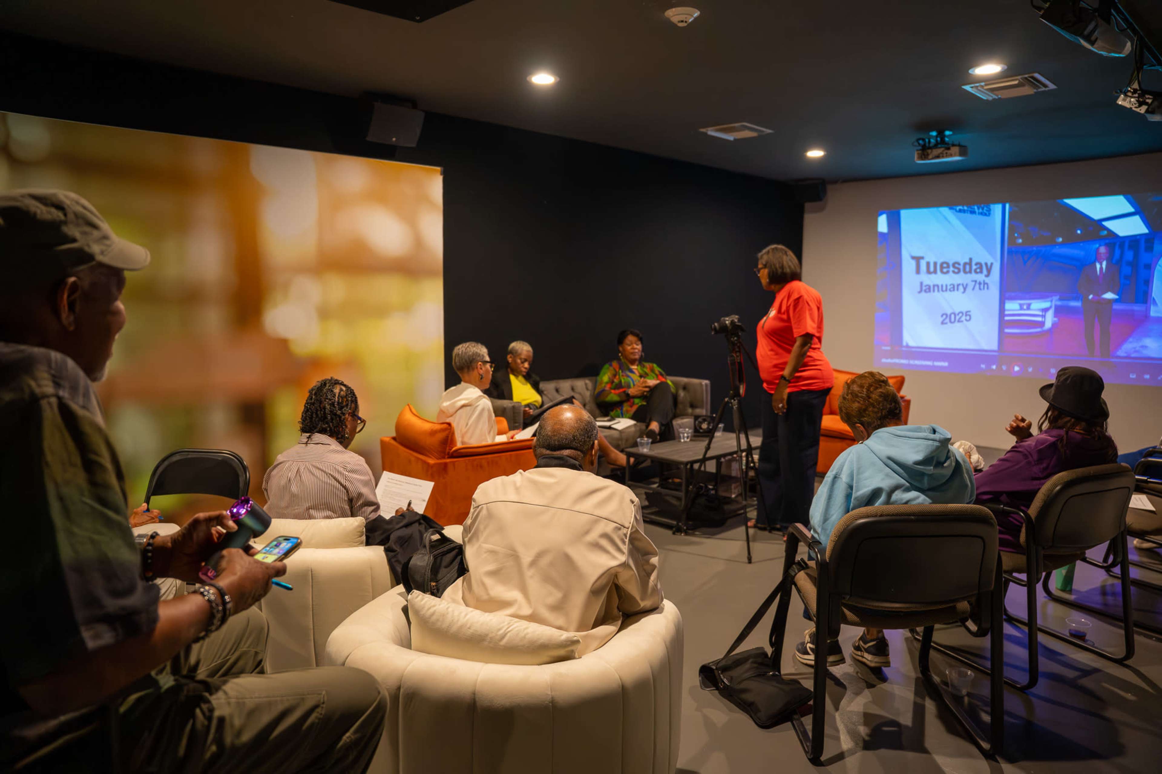 A group of people is seated in a small, modern room, while a woman in a red shirt speaks to them, and a screen displays "Tuesday, January 7, 2025."