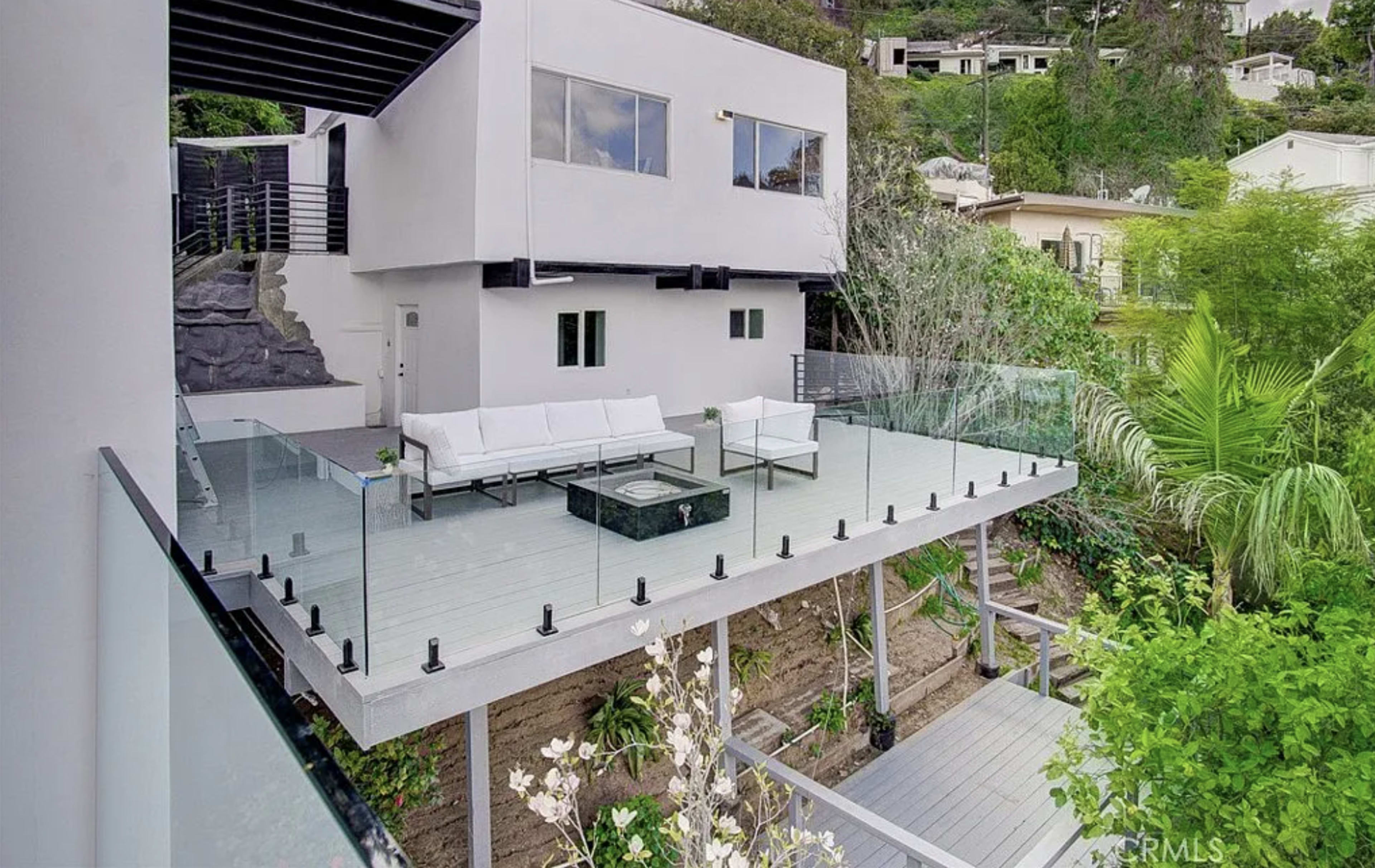 A modern house with large windows features a spacious deck surrounded by greenery and a glass railing.