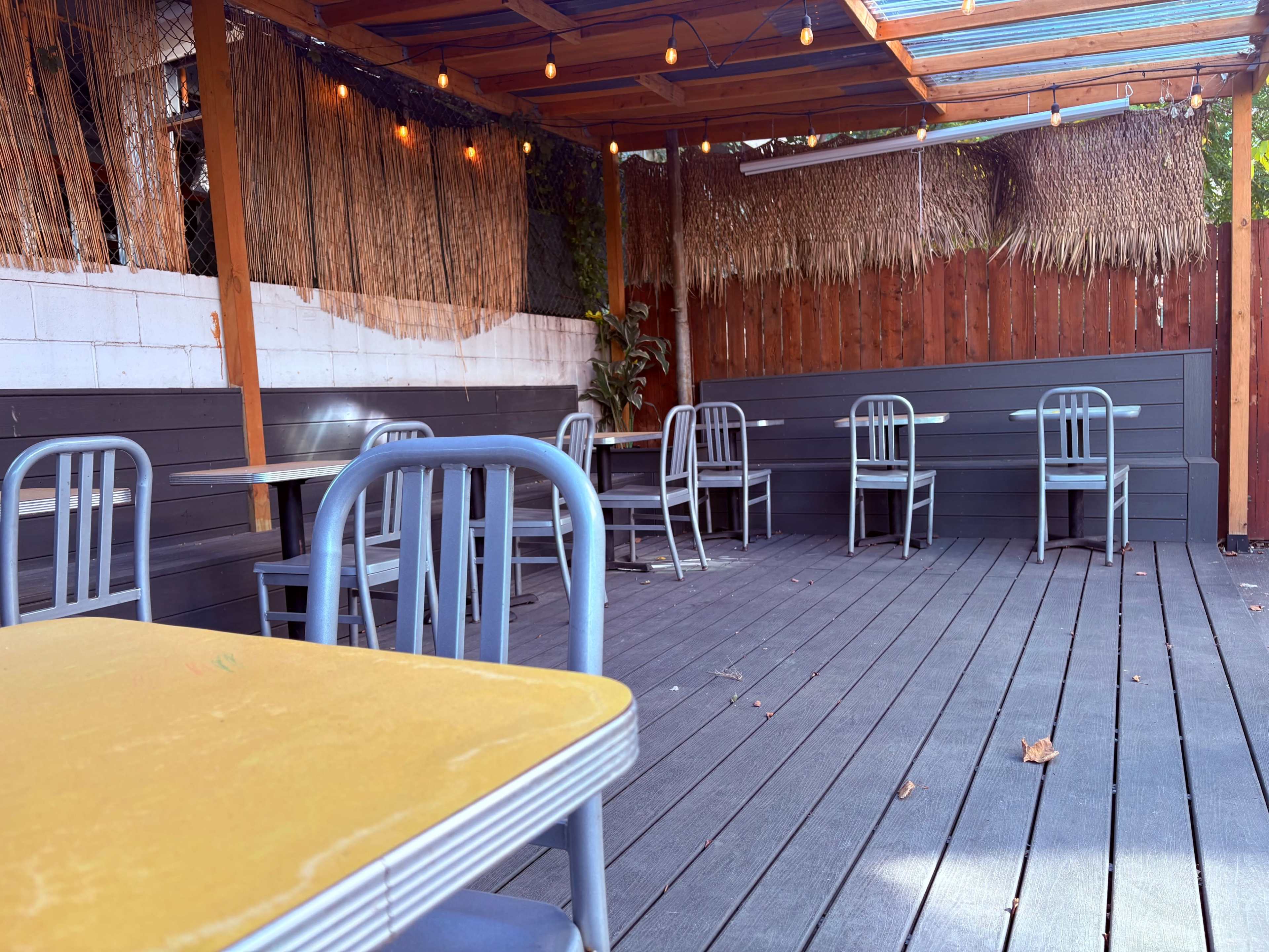 The image shows an outdoor seating area of a restaurant with wooden flooring, vacant tables and chairs, and overhead string lights.