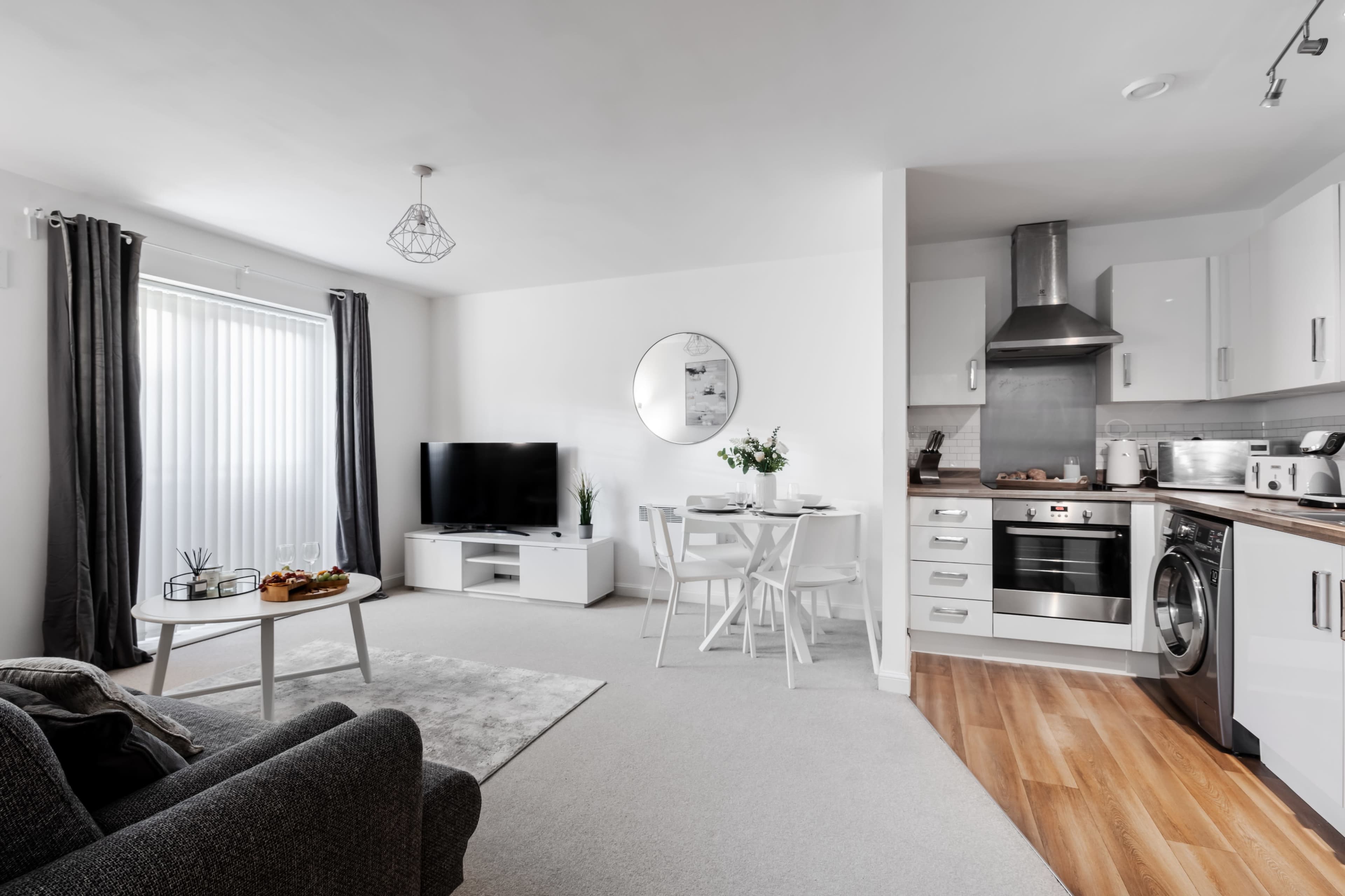 A modern living room and kitchen area with a television, a small dining table, and a gray sofa, featuring a neutral color palette and contemporary furnishings.