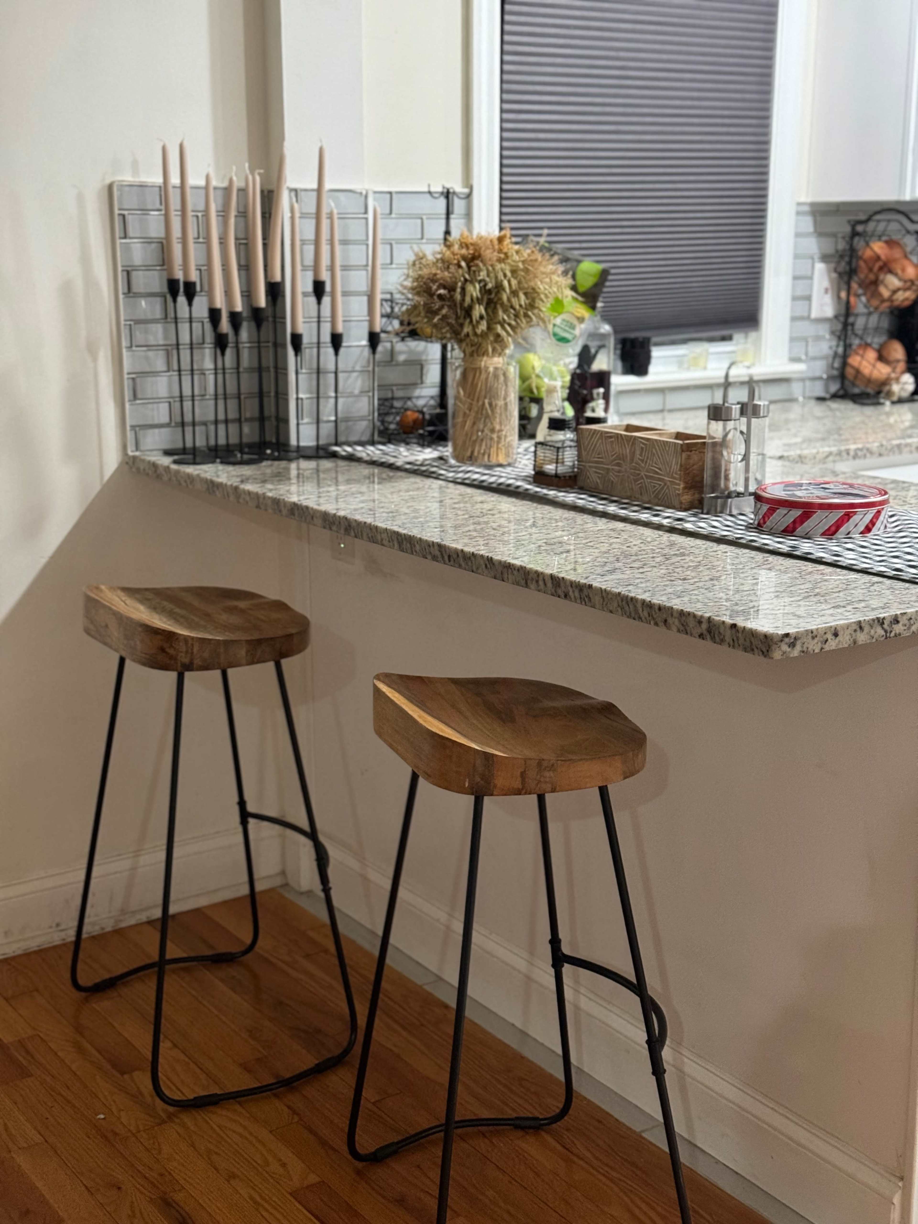 A kitchen bar area with two wooden stools, a granite countertop, and a decorative arrangement of candles and dried flowers in a glass vase.