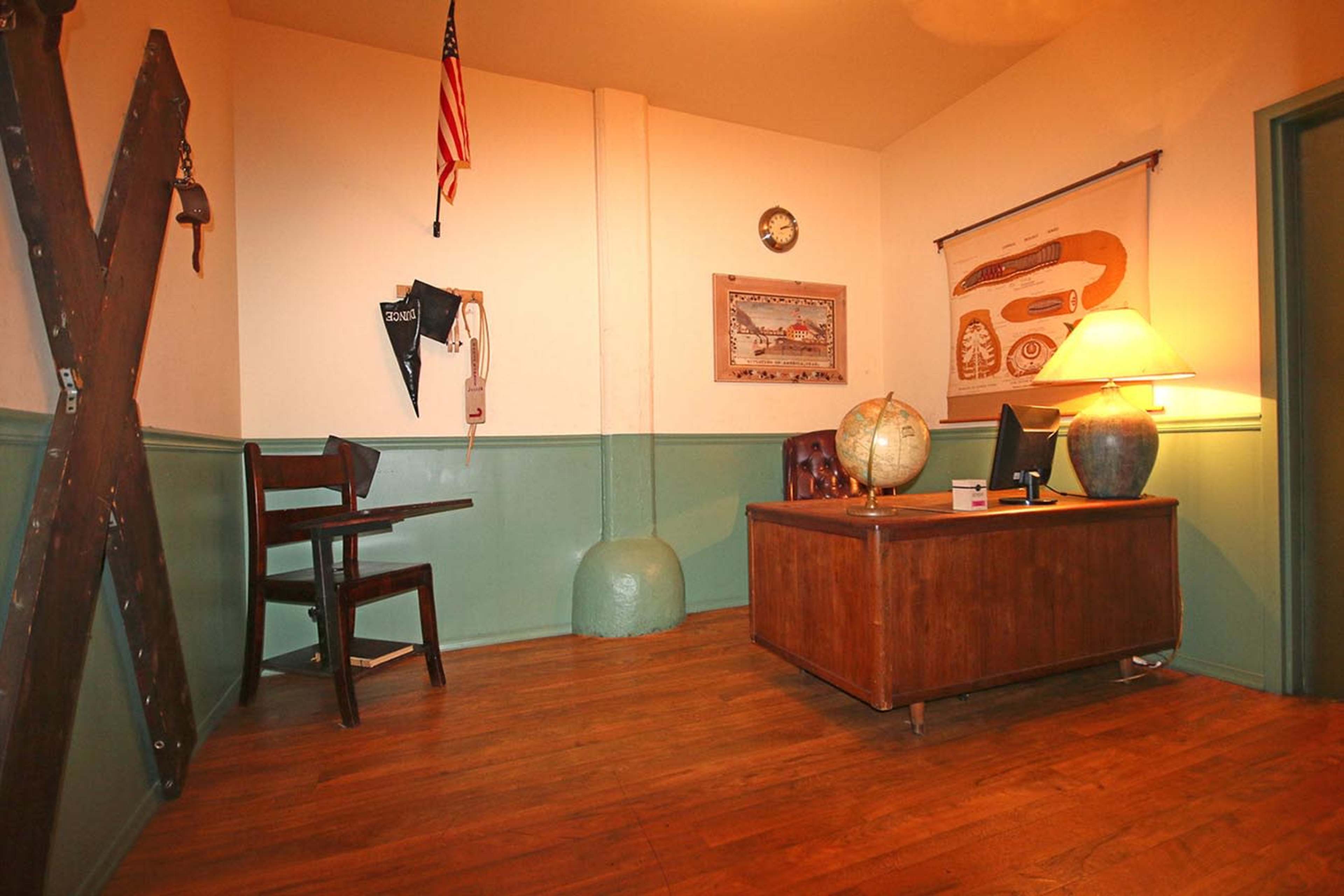 The image shows a sparsely furnished office with a wooden desk, a globe, a chair, an American flag, and a clock on the wall.