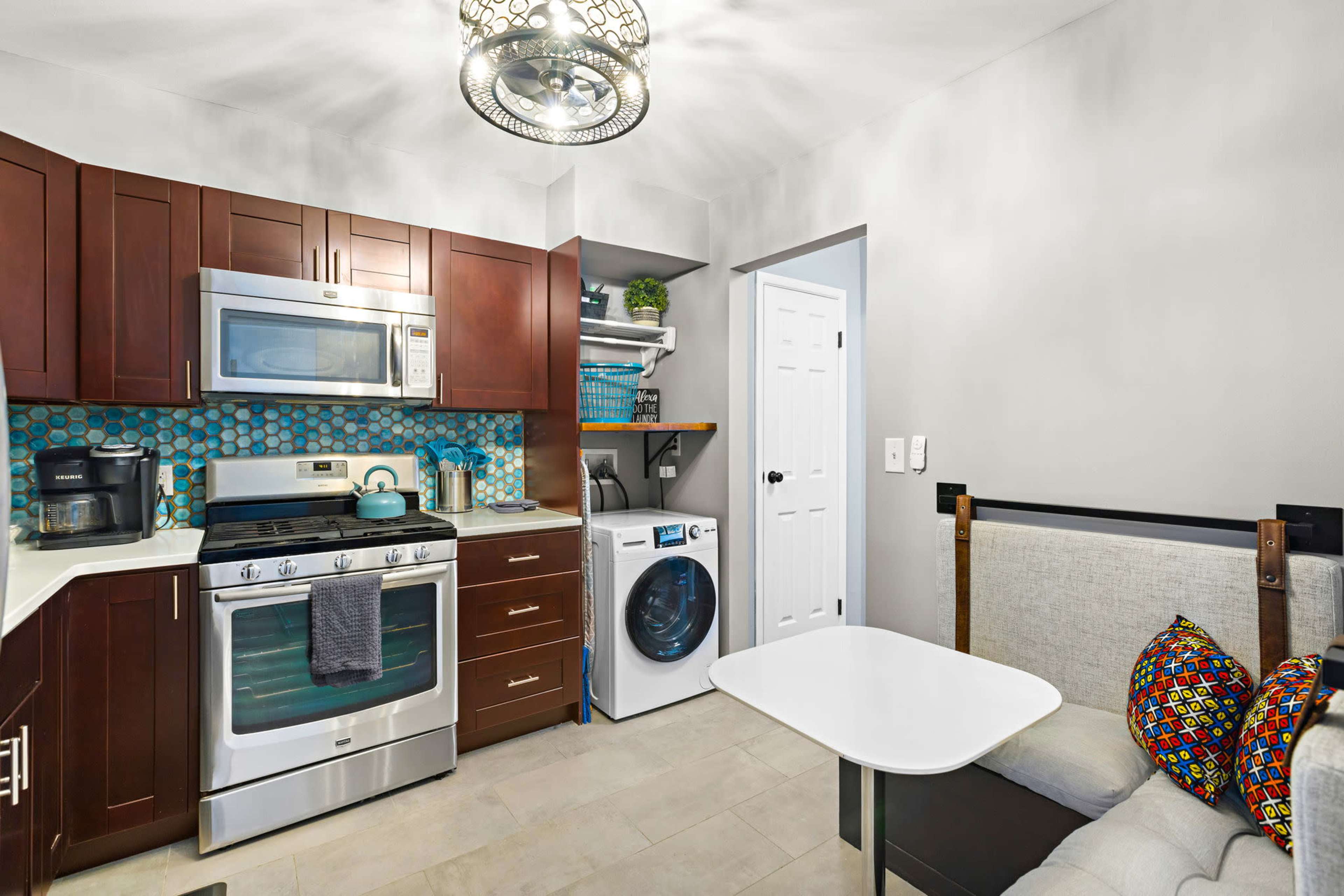 The kitchen features dark wooden cabinets, stainless steel appliances, and a small table with decorative pillows beside a washer and dryer.