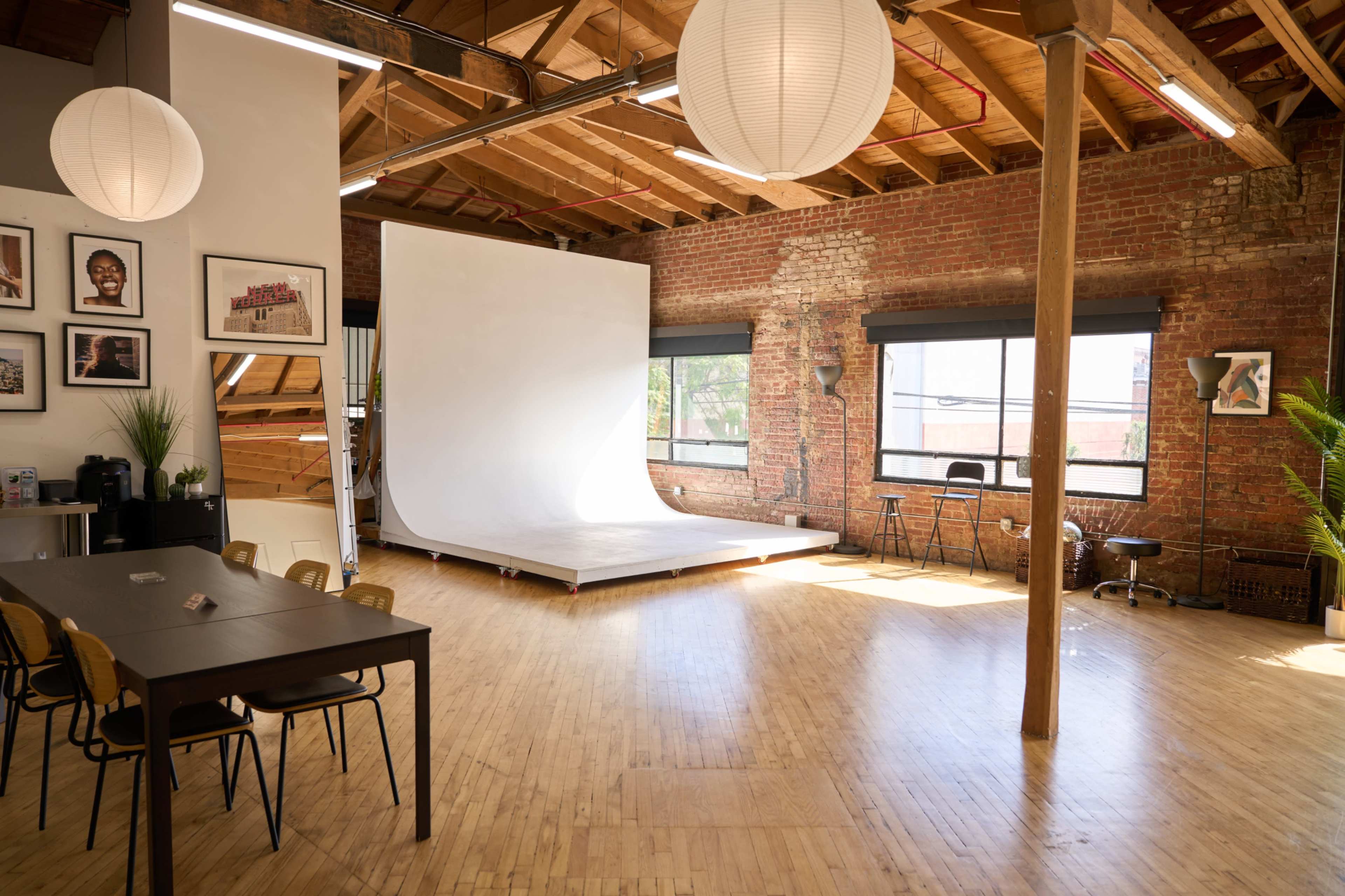 The image shows a spacious studio with a wooden floor, exposed brick walls, and a large white backdrop, accompanied by a dining area featuring a table and chairs.