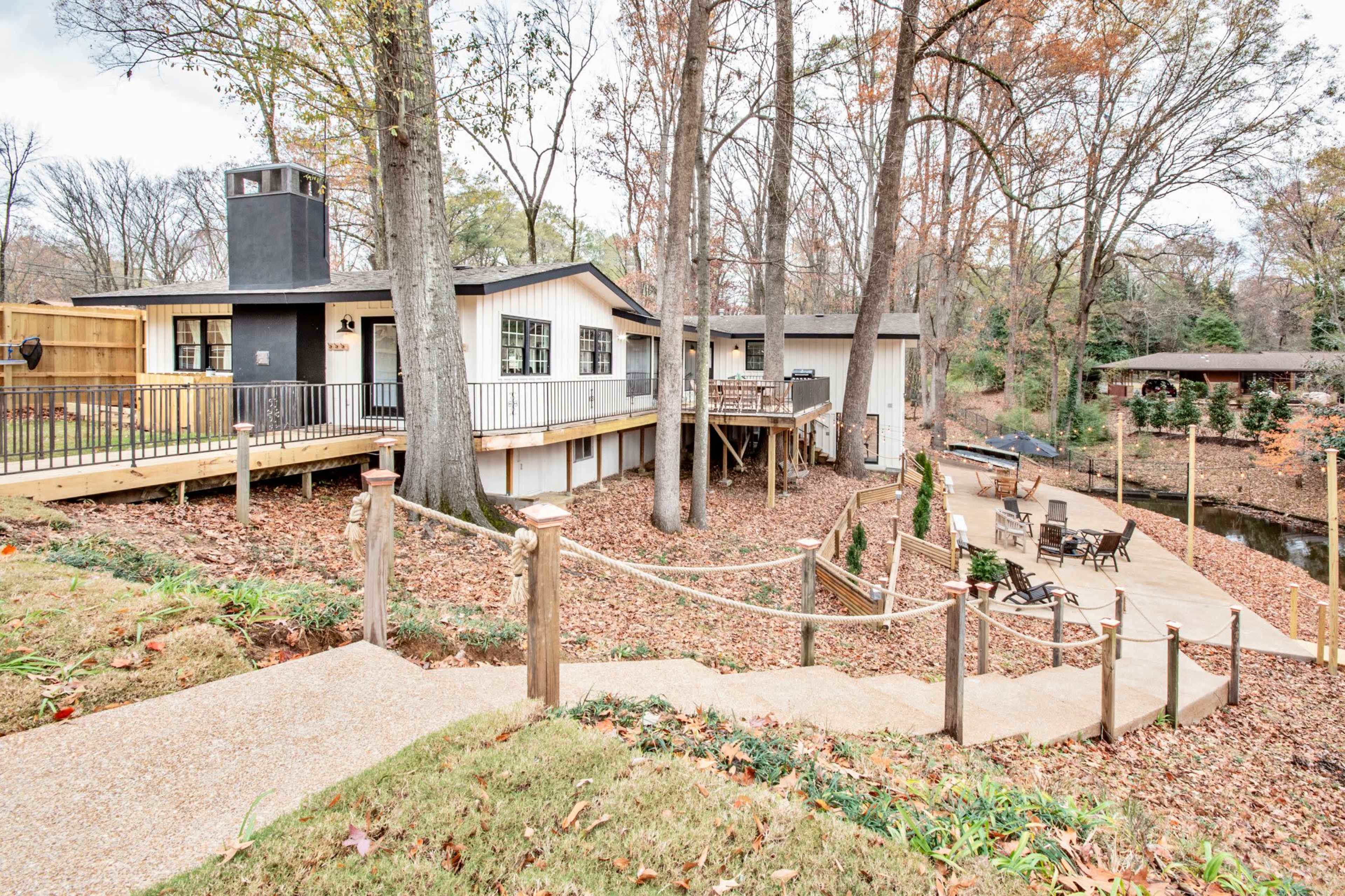 A modern-looking house with a patio and a wooden deck is surrounded by trees and autumn leaves, with a pathway leading down to a dock by a small body of water.