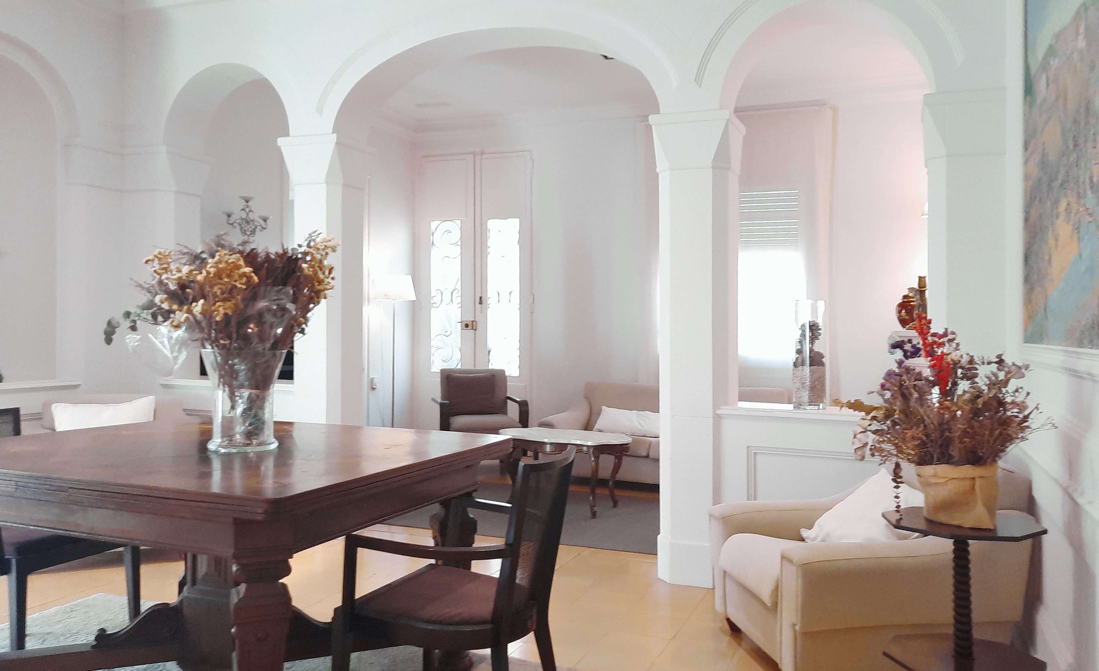 The image shows a bright living room featuring a wooden dining table surrounded by chairs, with decorative vases of dried flowers placed around the space.