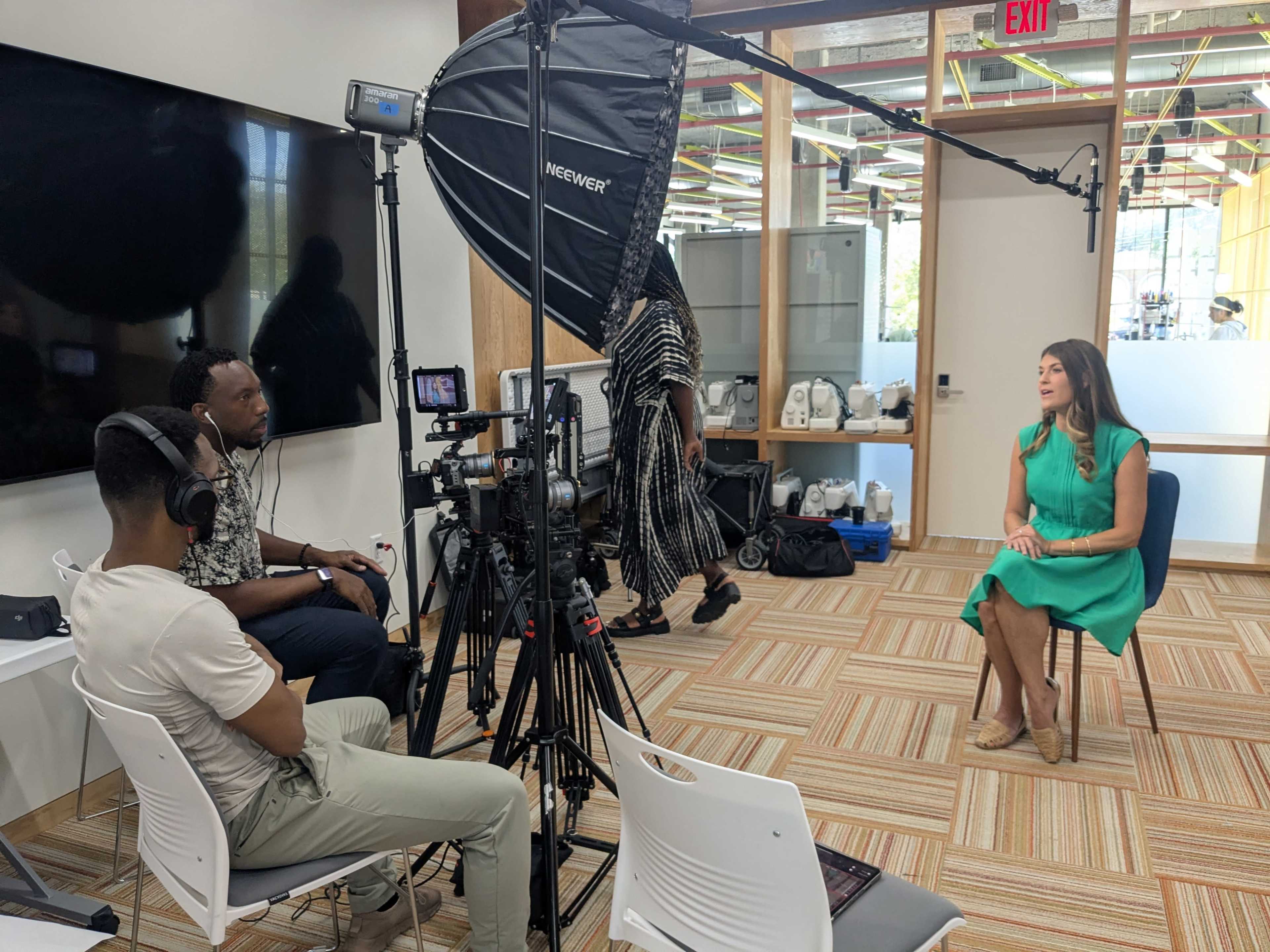 A woman in a green dress is seated for an interview in a well-lit office with a camera and lighting setup, while two men are adjusting equipment nearby.