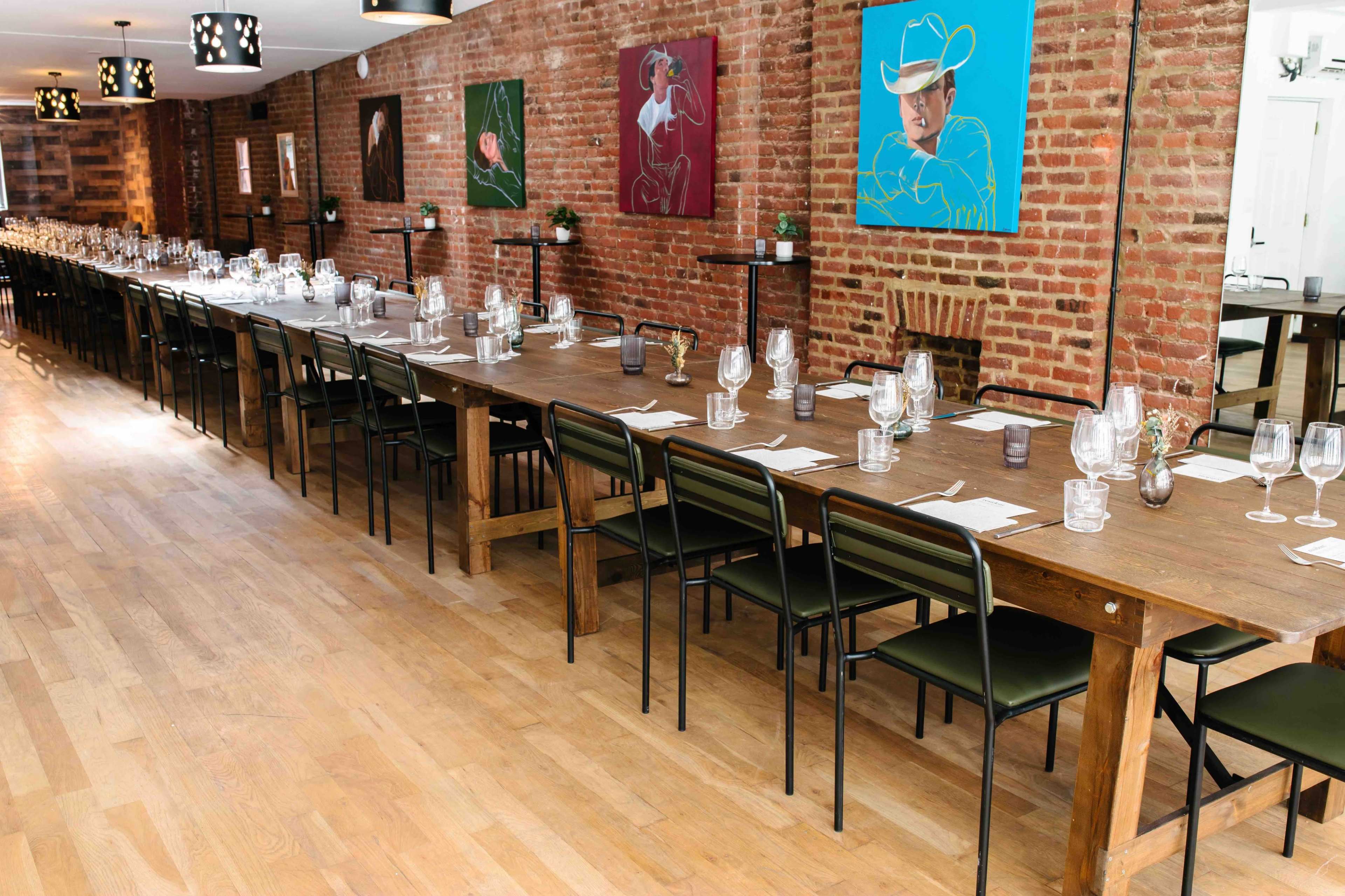 A long wooden dining table is set with place settings and glassware, surrounded by black chairs, in a space featuring exposed brick walls and colorful artworks.
