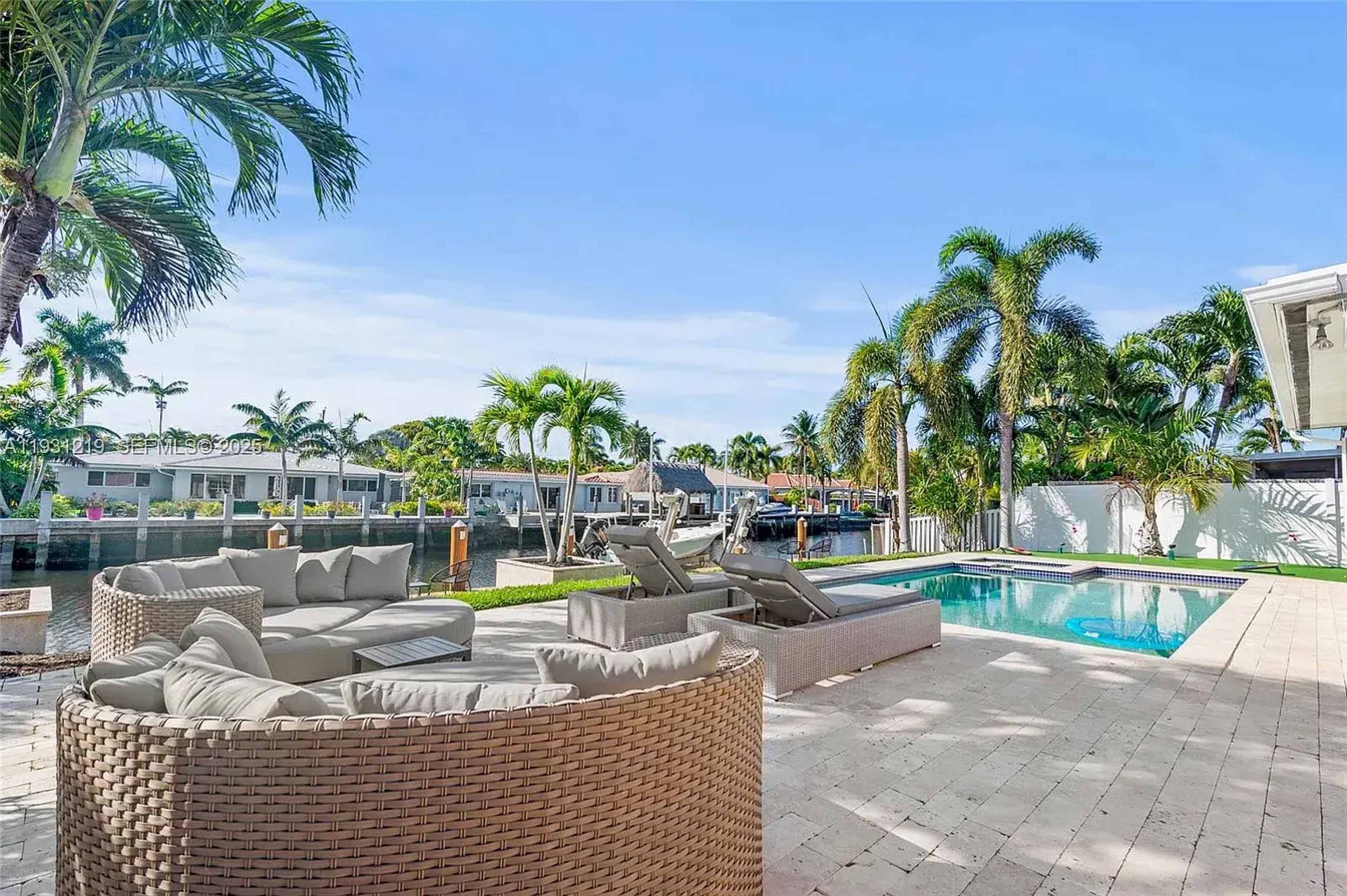 The image shows a backyard with a swimming pool, lounge chairs, and a seating area, all surrounded by palm trees and a waterfront view.