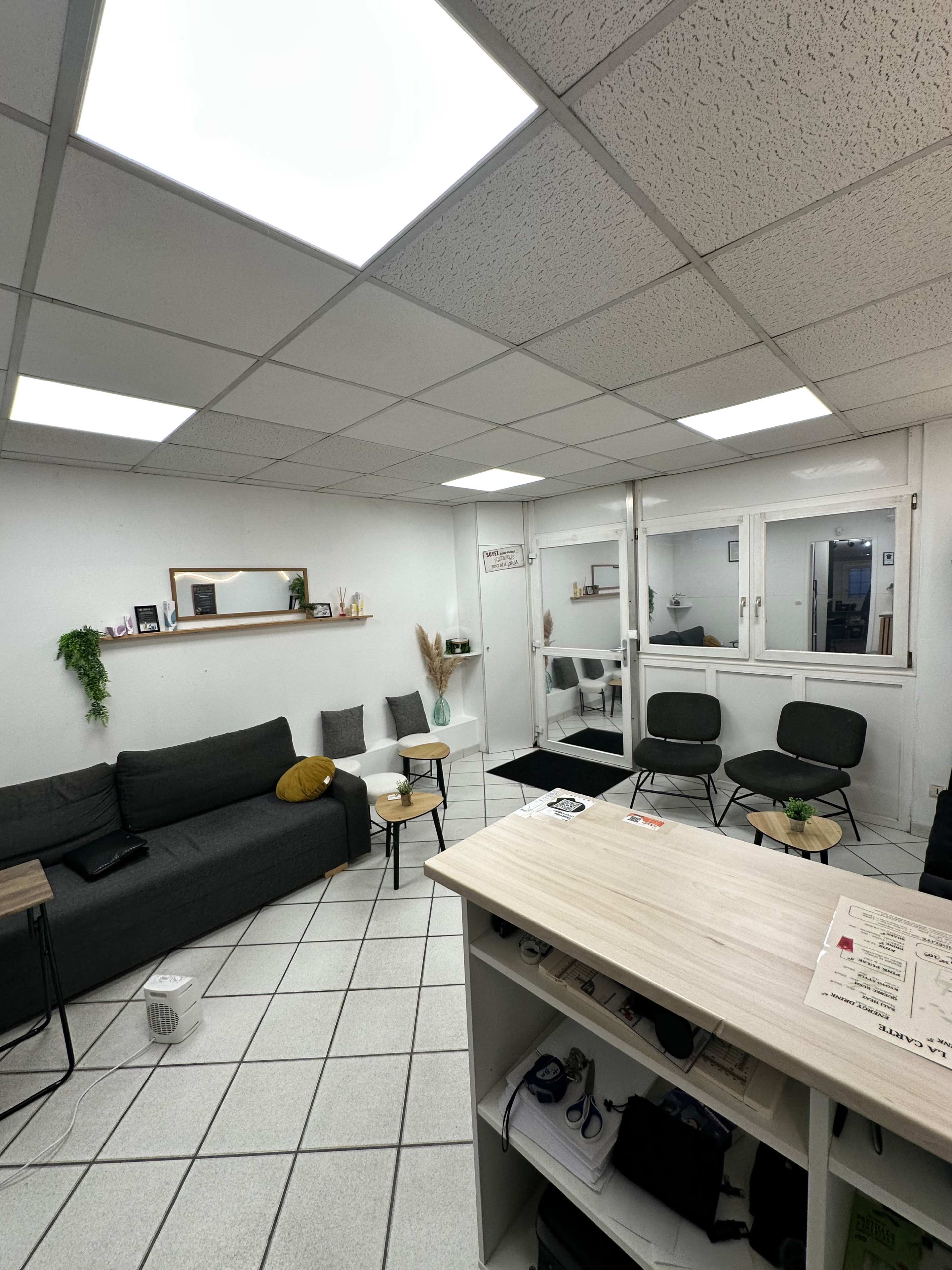 The image shows a modern office space with a seating area, a small table, and large windows that provide natural light.