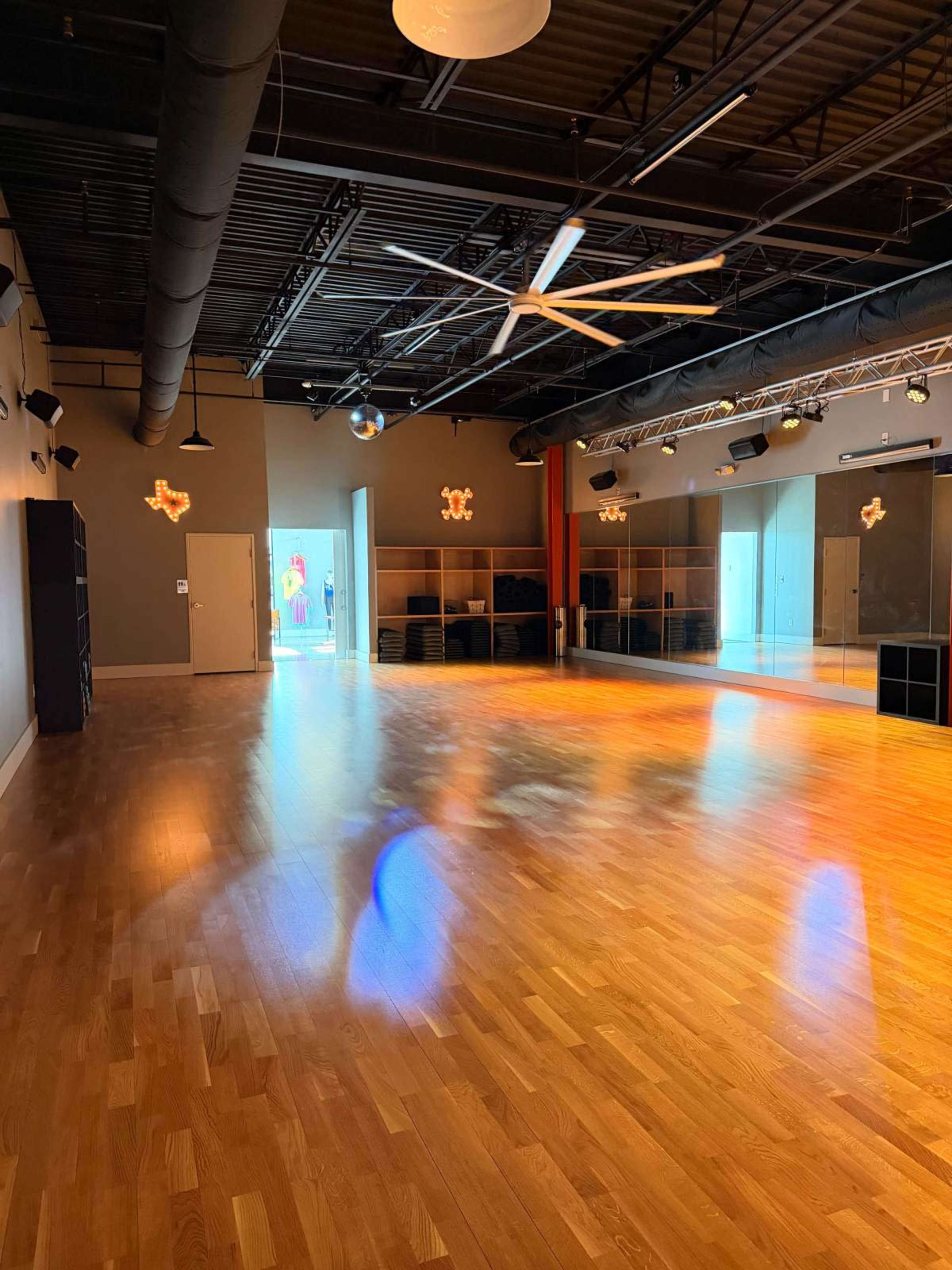 The image shows an empty dance studio featuring wooden flooring, large mirrors on one wall, and a bright light source illuminating the space.