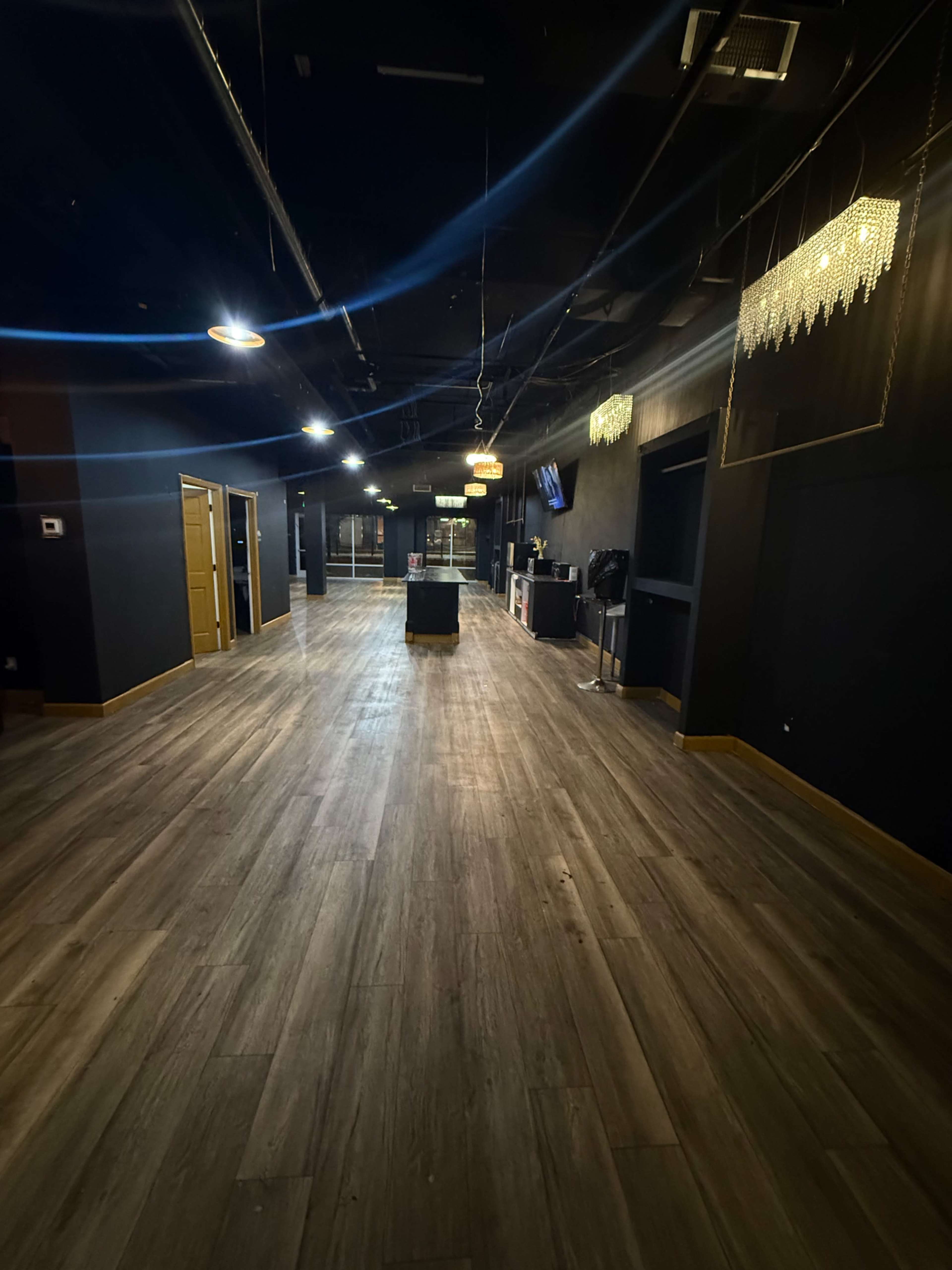 The image shows a spacious, dimly lit interior with wooden flooring, featuring a long corridor leading to various rooms and hanging light fixtures.