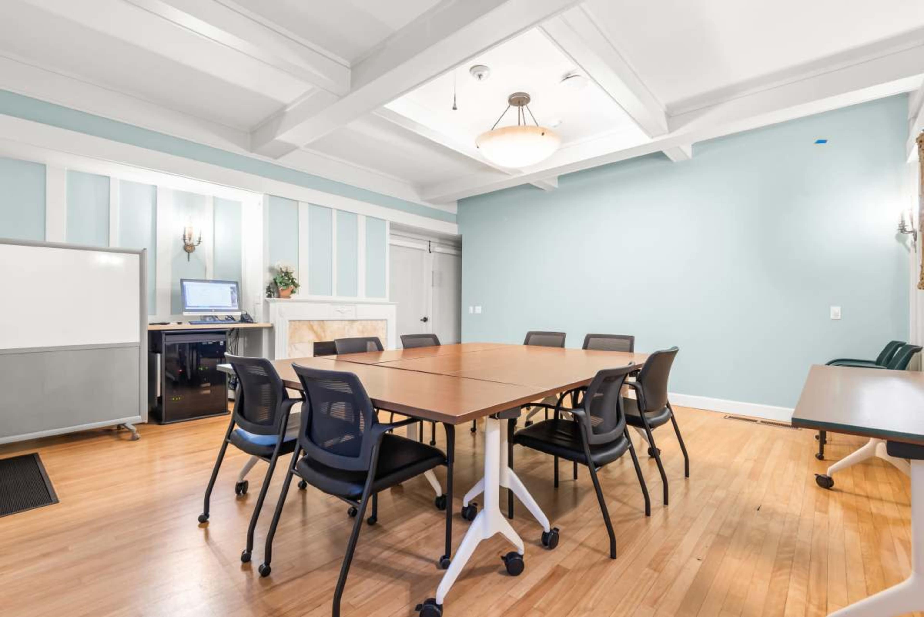 A spacious meeting room features a large table surrounded by black chairs, with a whiteboard and a computer station against one wall.