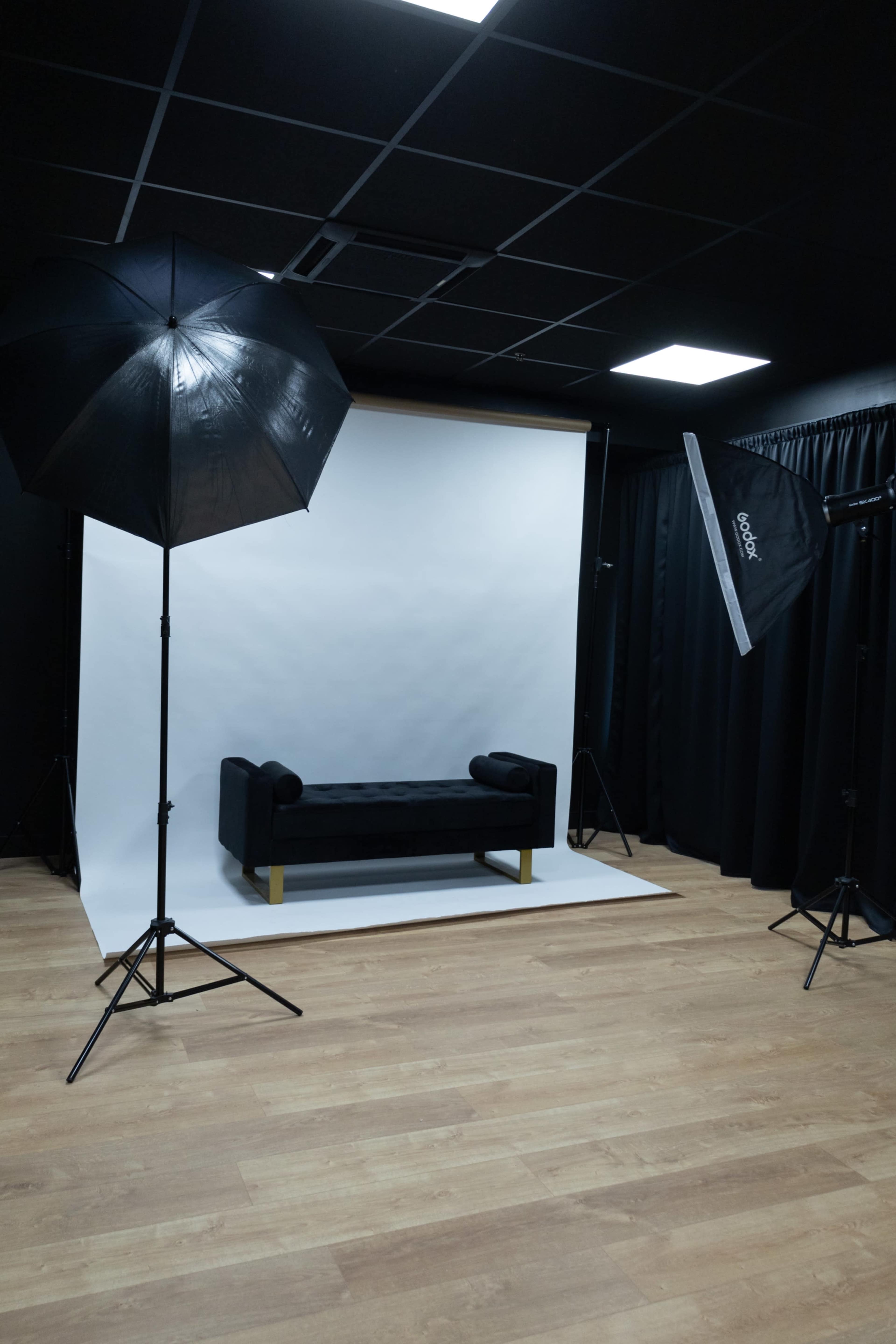A photography studio features a black couch centered against a white backdrop, illuminated by two softbox lights.