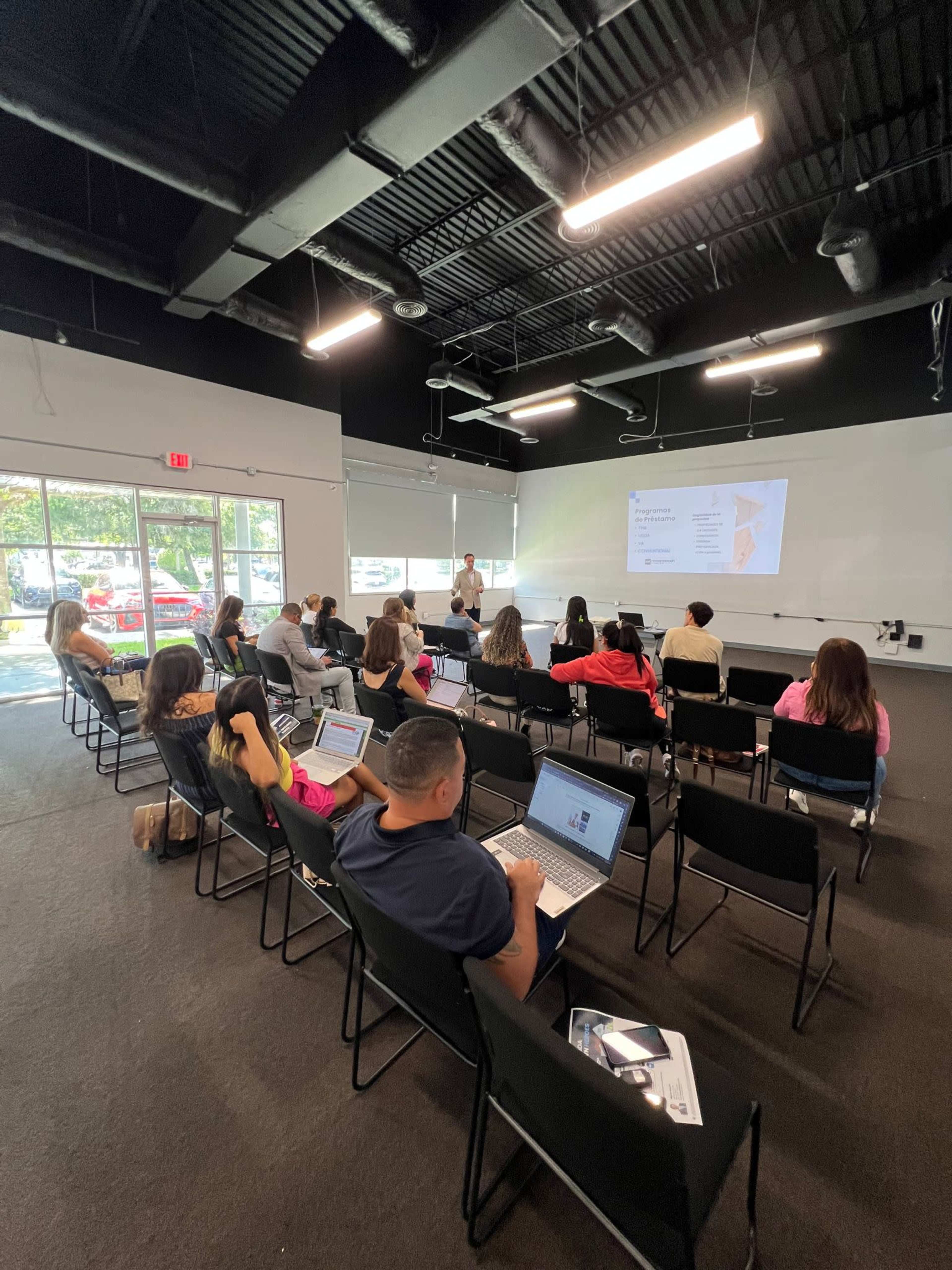 A speaker is presenting in a classroom setting where attendees are seated and working on laptops.