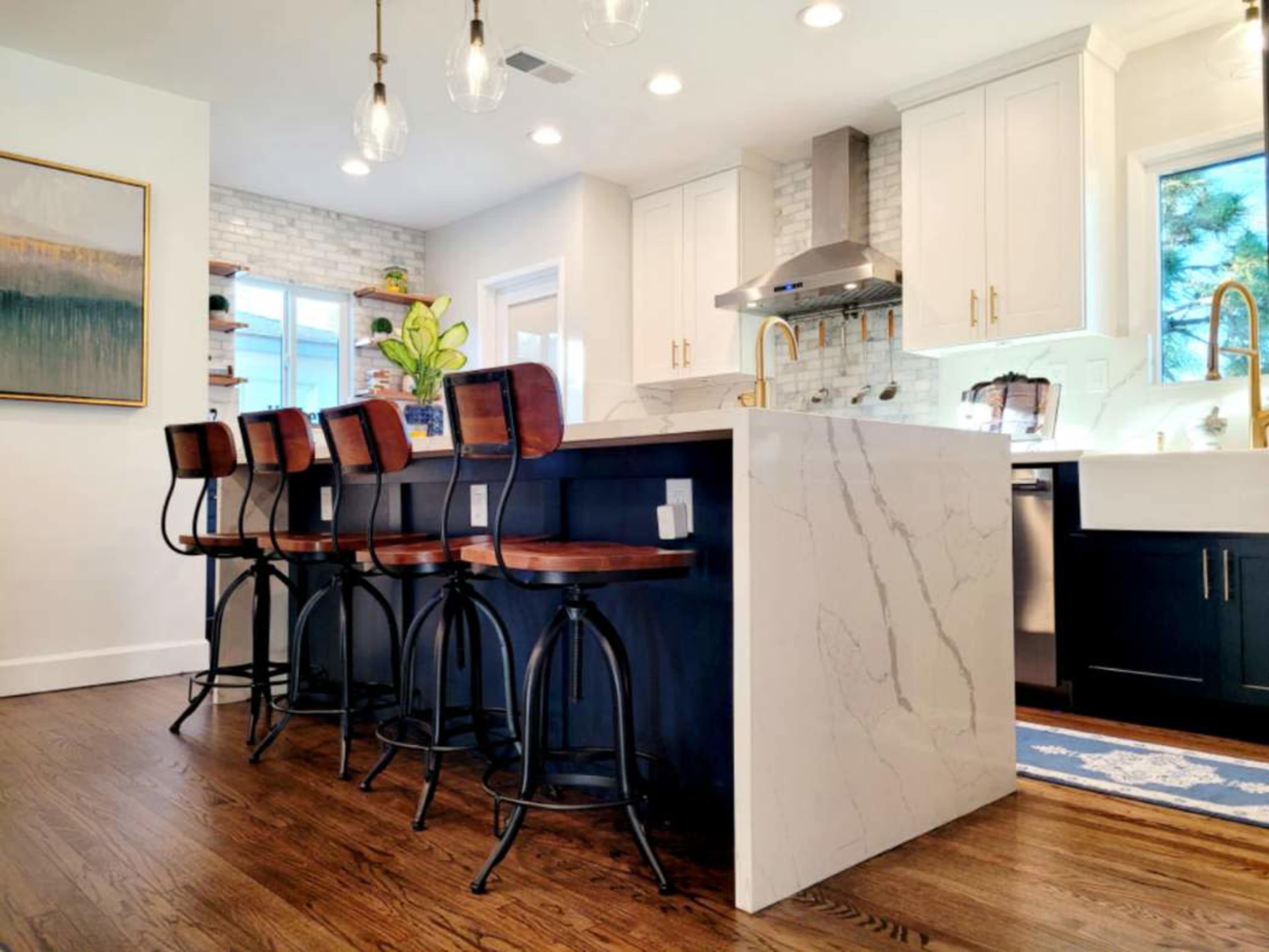 A modern kitchen featuring a large island with a marble countertop and four wooden bar stools, along with white cabinets and stainless steel appliances.