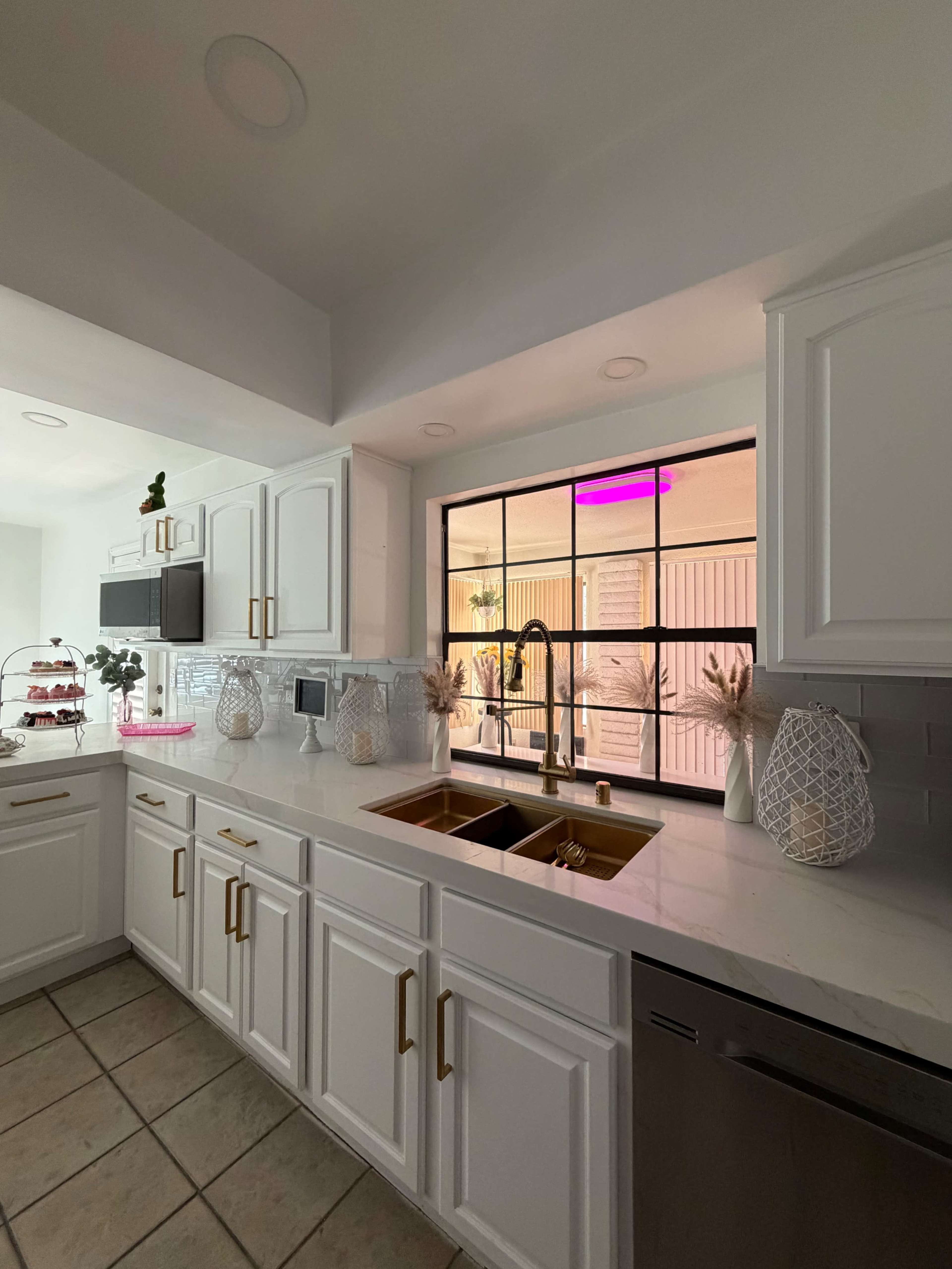 The image shows a modern kitchen with white cabinets, a double sink, and a large window overlooking a patio with minimal decor.