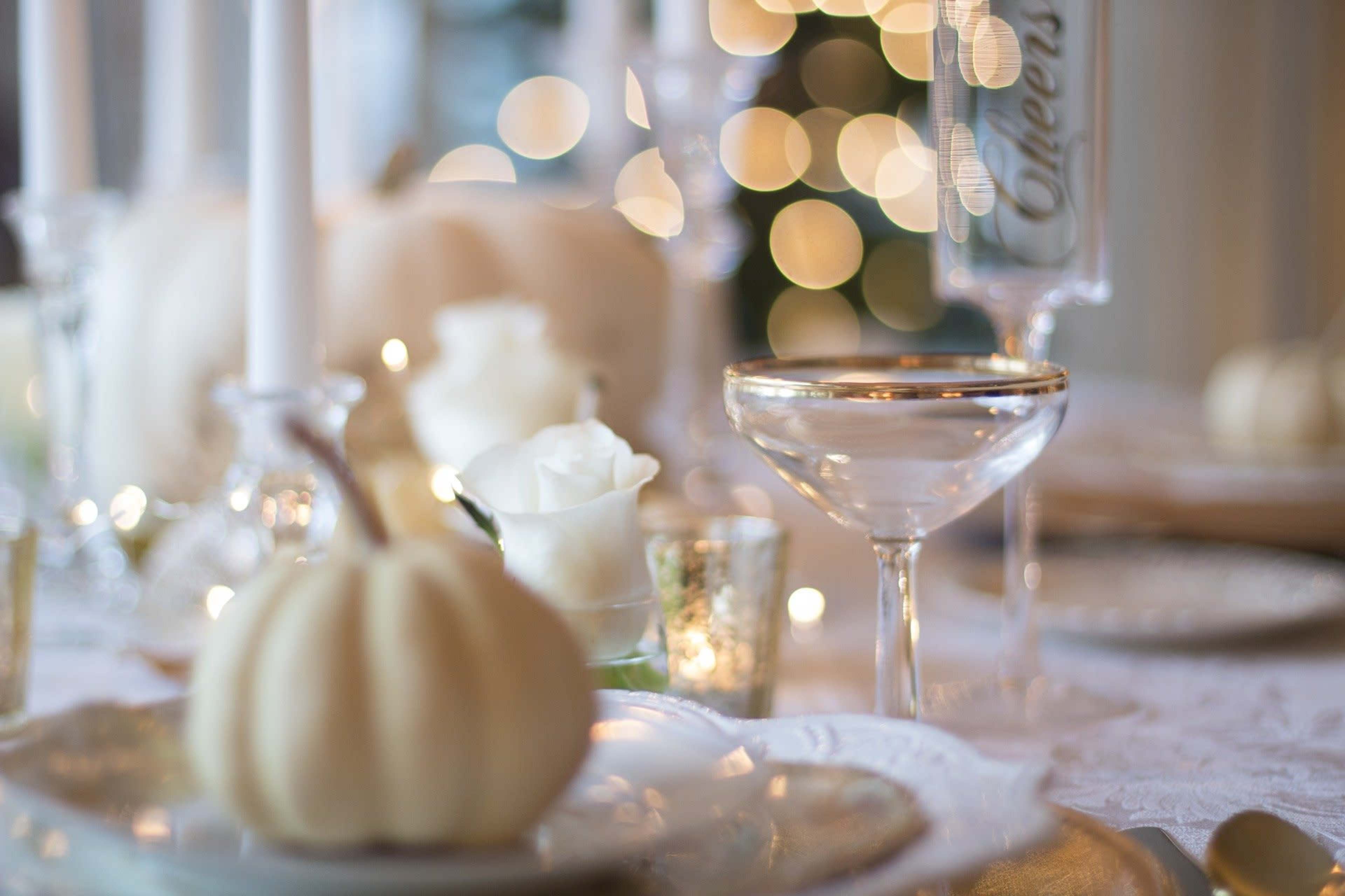 A elegantly set dining table features white pumpkins, roses, and sparkling glasses beside soft, blurred lights in the background.