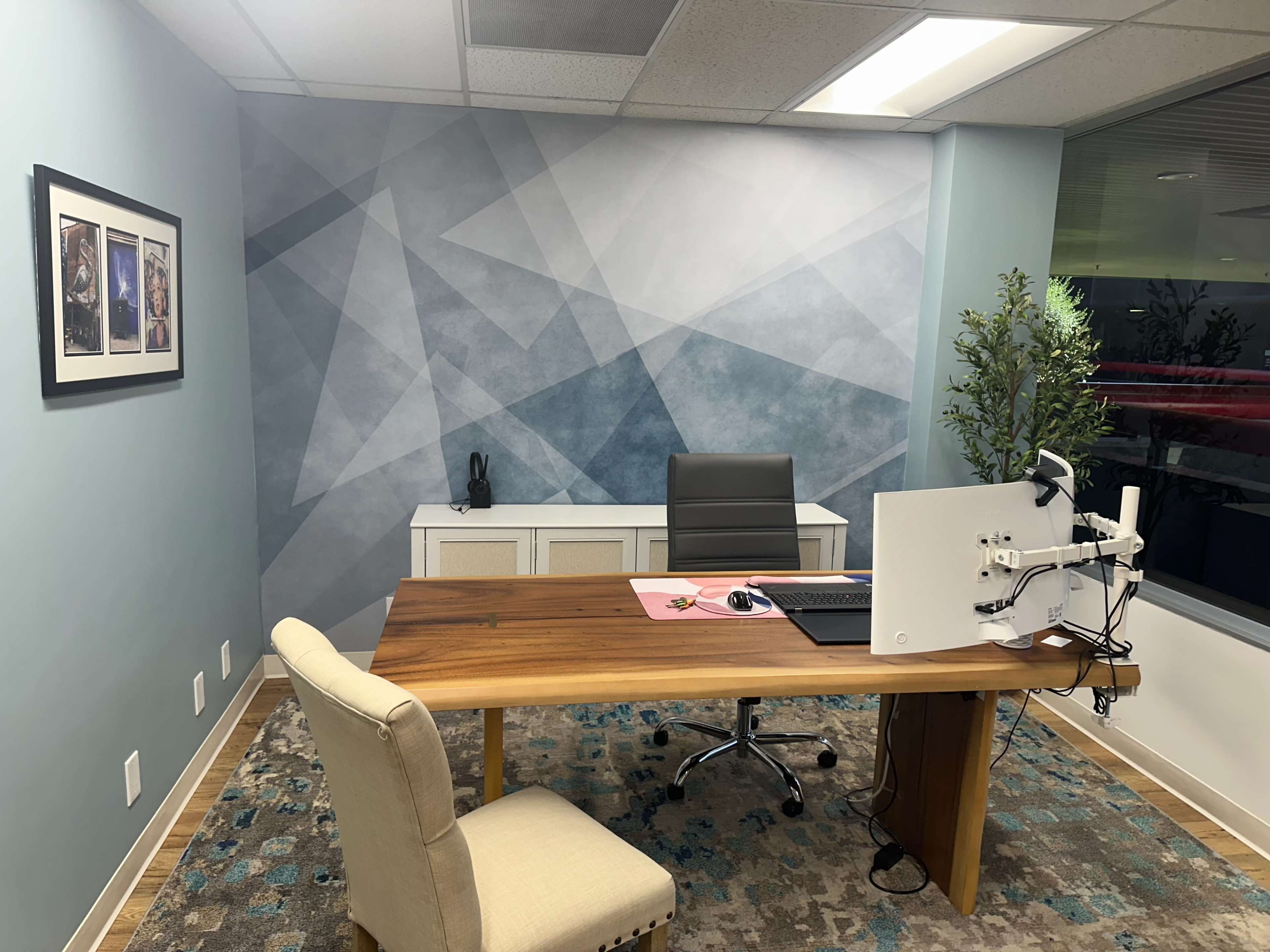 A modern office space featuring a wooden desk, ergonomic chair, computer monitor on an arm, and a decorative wall with geometric patterns.