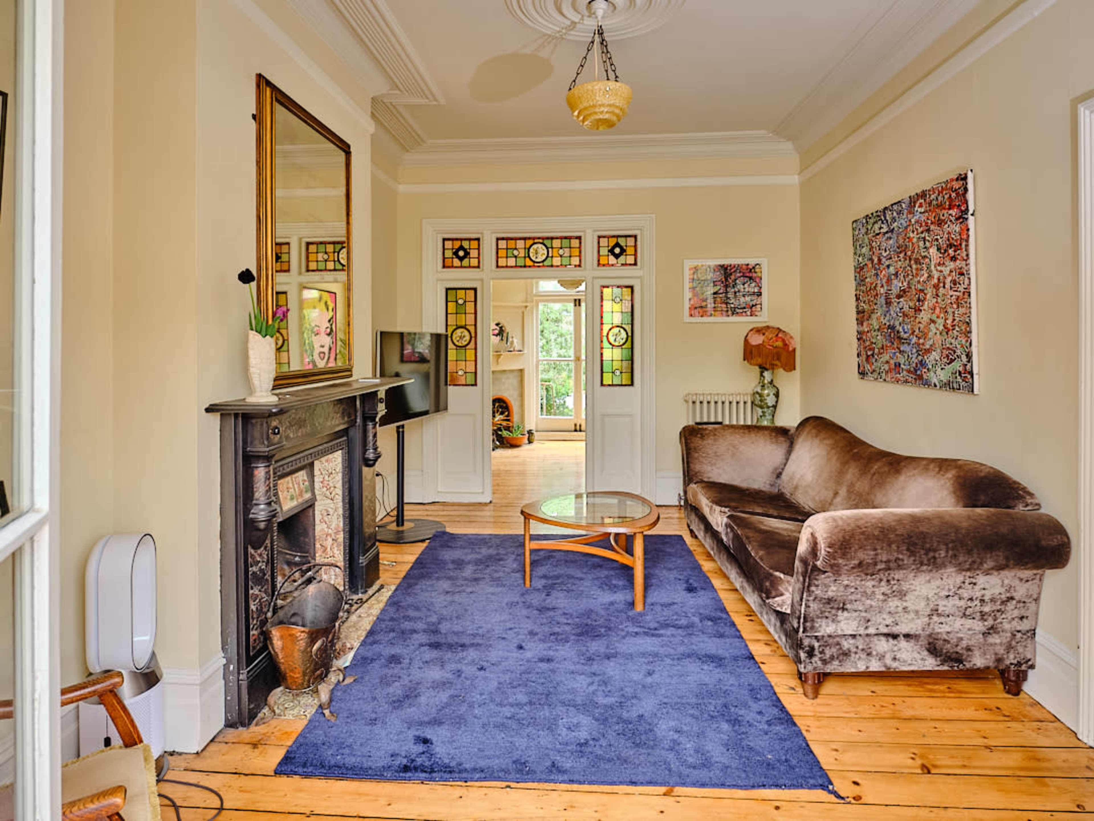 The image shows a living room with a brown plush couch, a glass coffee table on a blue rug, and a decorative fireplace with a large mirror above it.