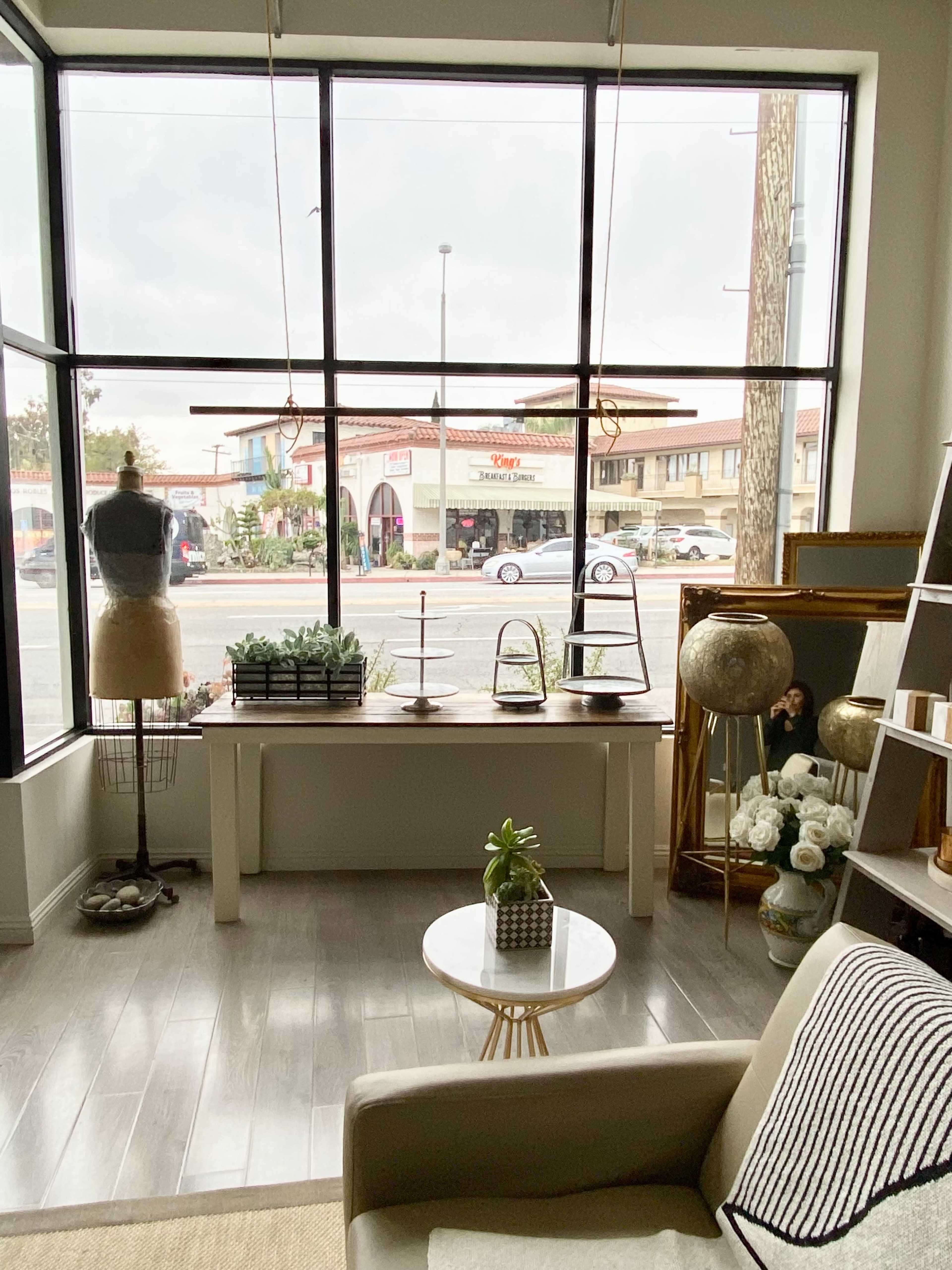A modern interior space featuring a table with decorative items, a mannequin, and large windows overlooking a street with shops outside.