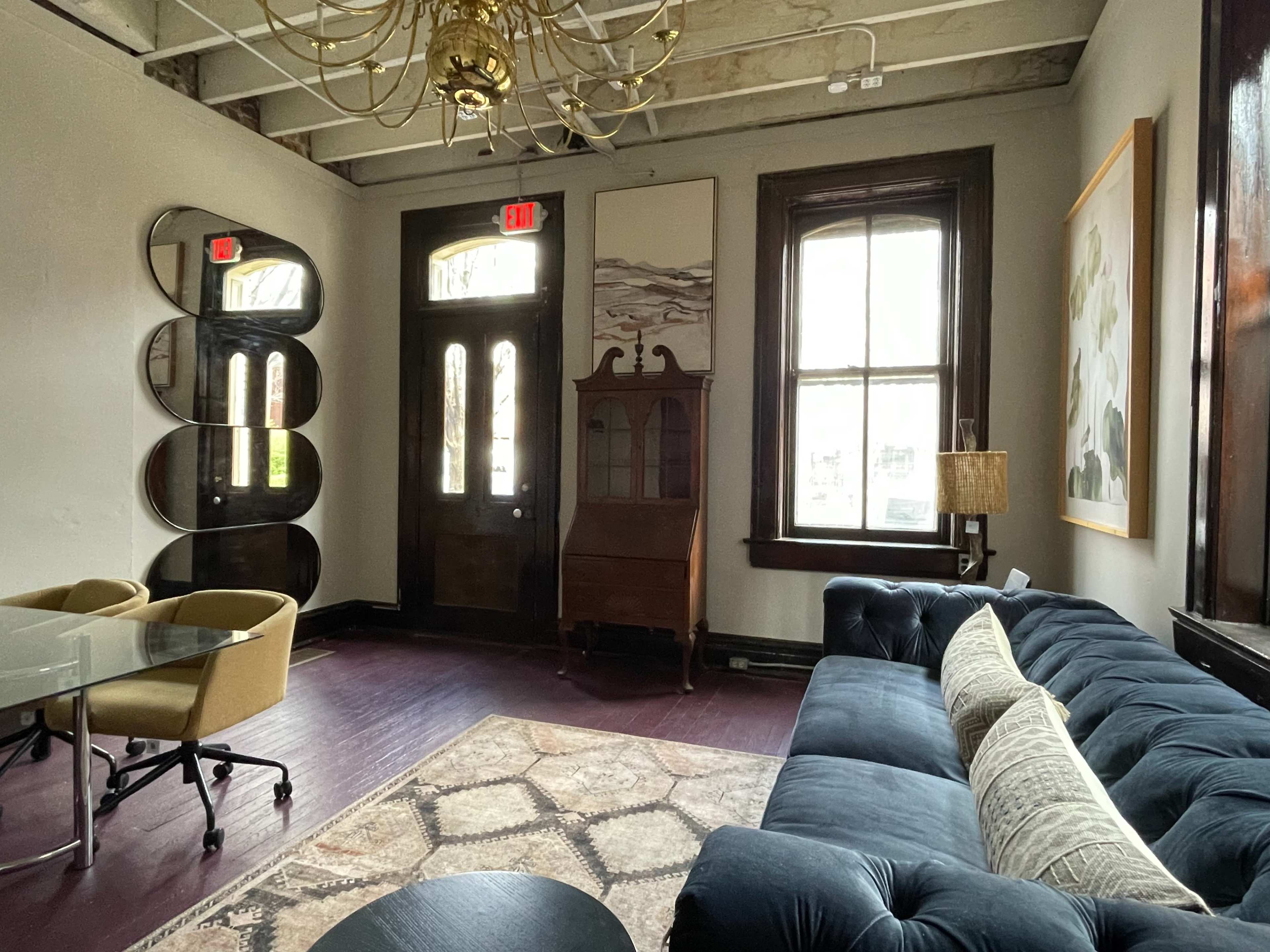 A stylish lounge area with a dark blue sofa, a glass table, and a wooden cabinet, illuminated by natural light coming through large windows.