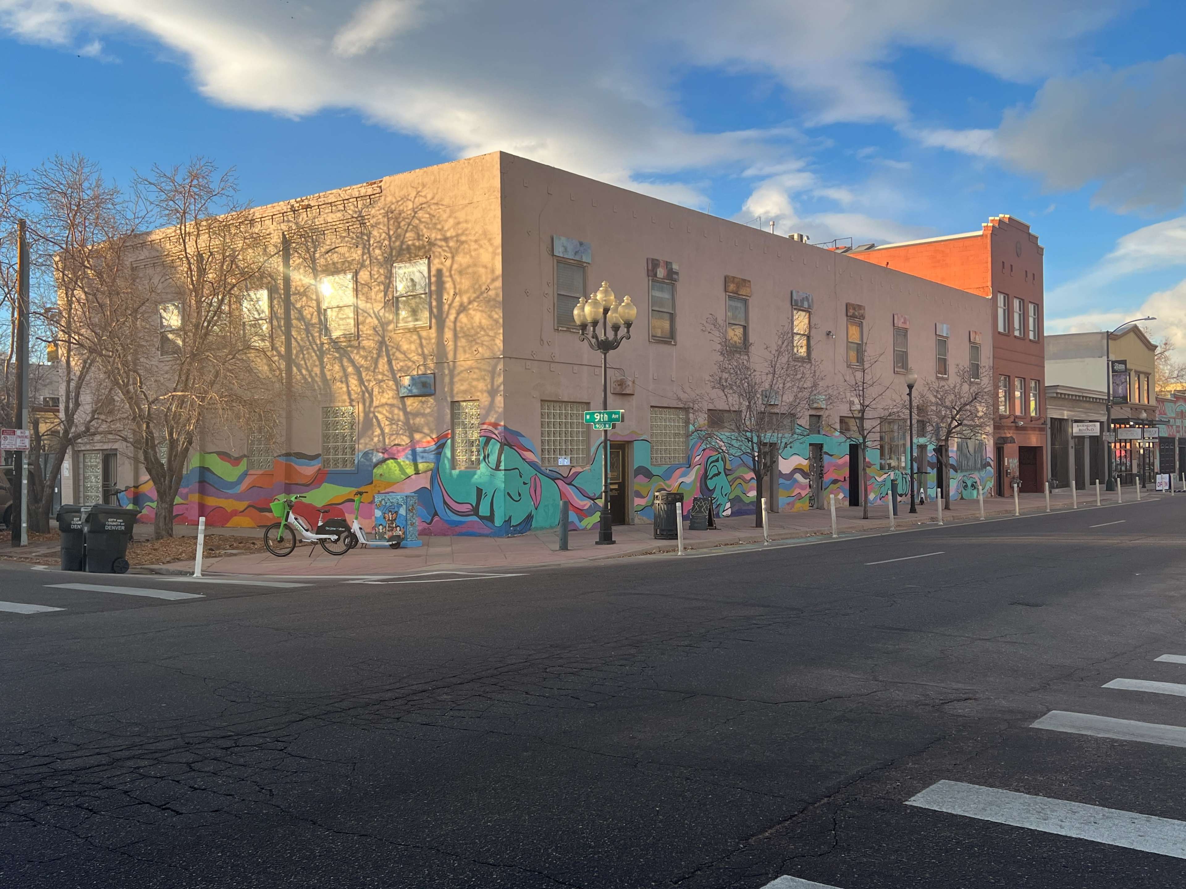 The image shows a colorful mural spanning the side of a building on a city street, with several bare trees and parked bicycles nearby.