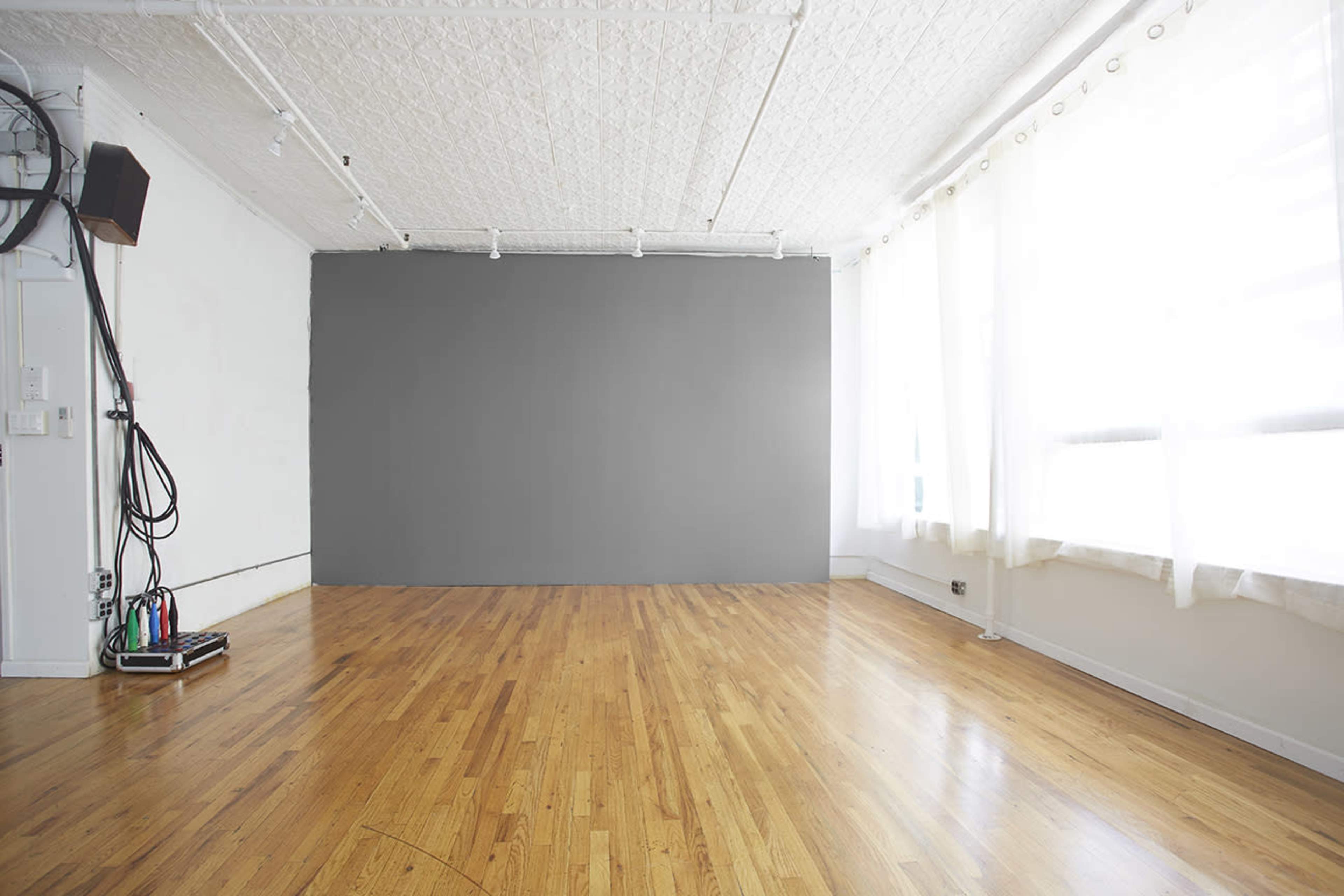 The image shows a spacious room with hardwood floors, large windows dressed in white curtains, and a gray wall at one end.