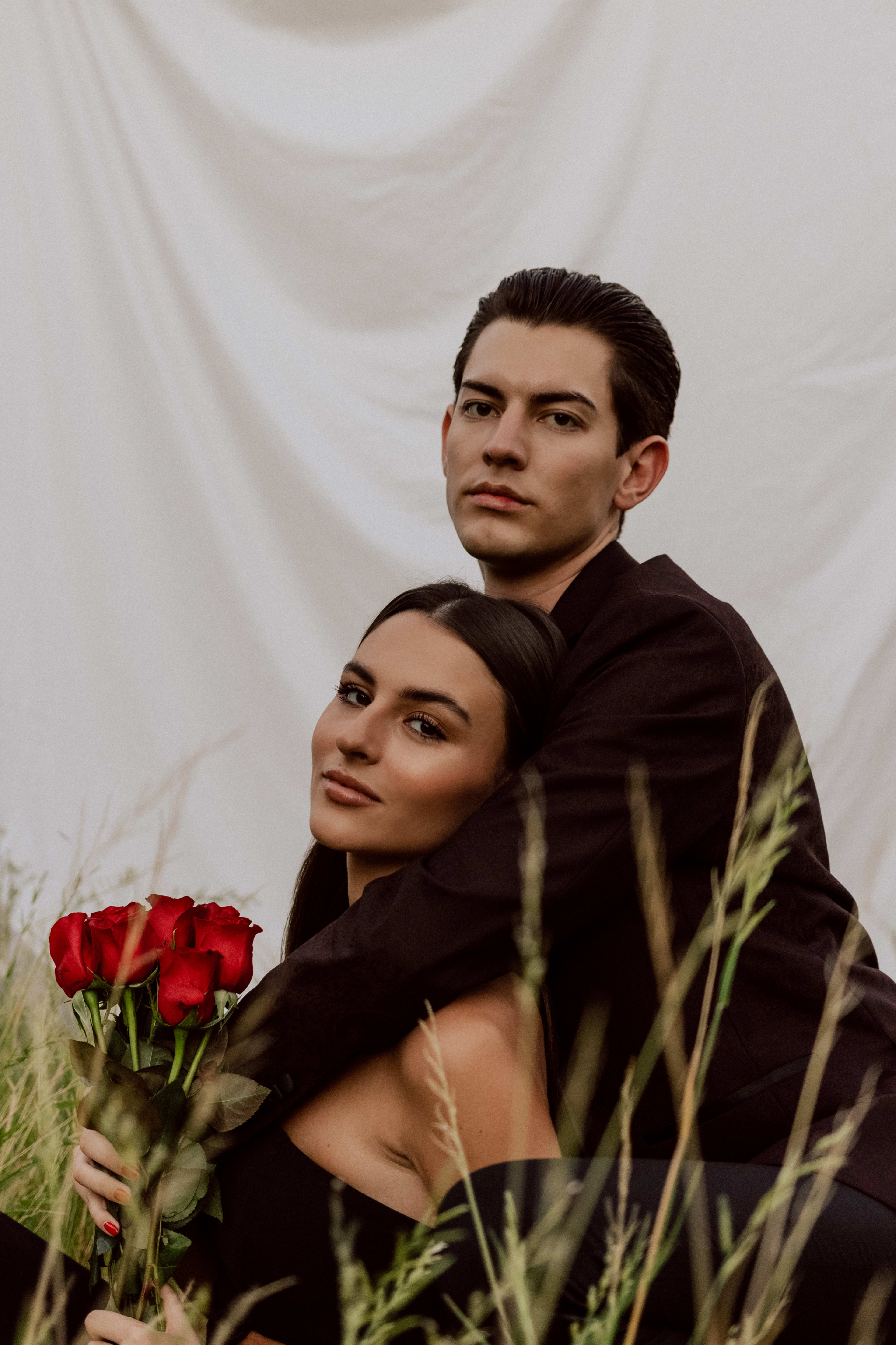 A couple holding red roses poses in beige clothing for an outdoor photo shoot in a garden surrounded by tall grass.