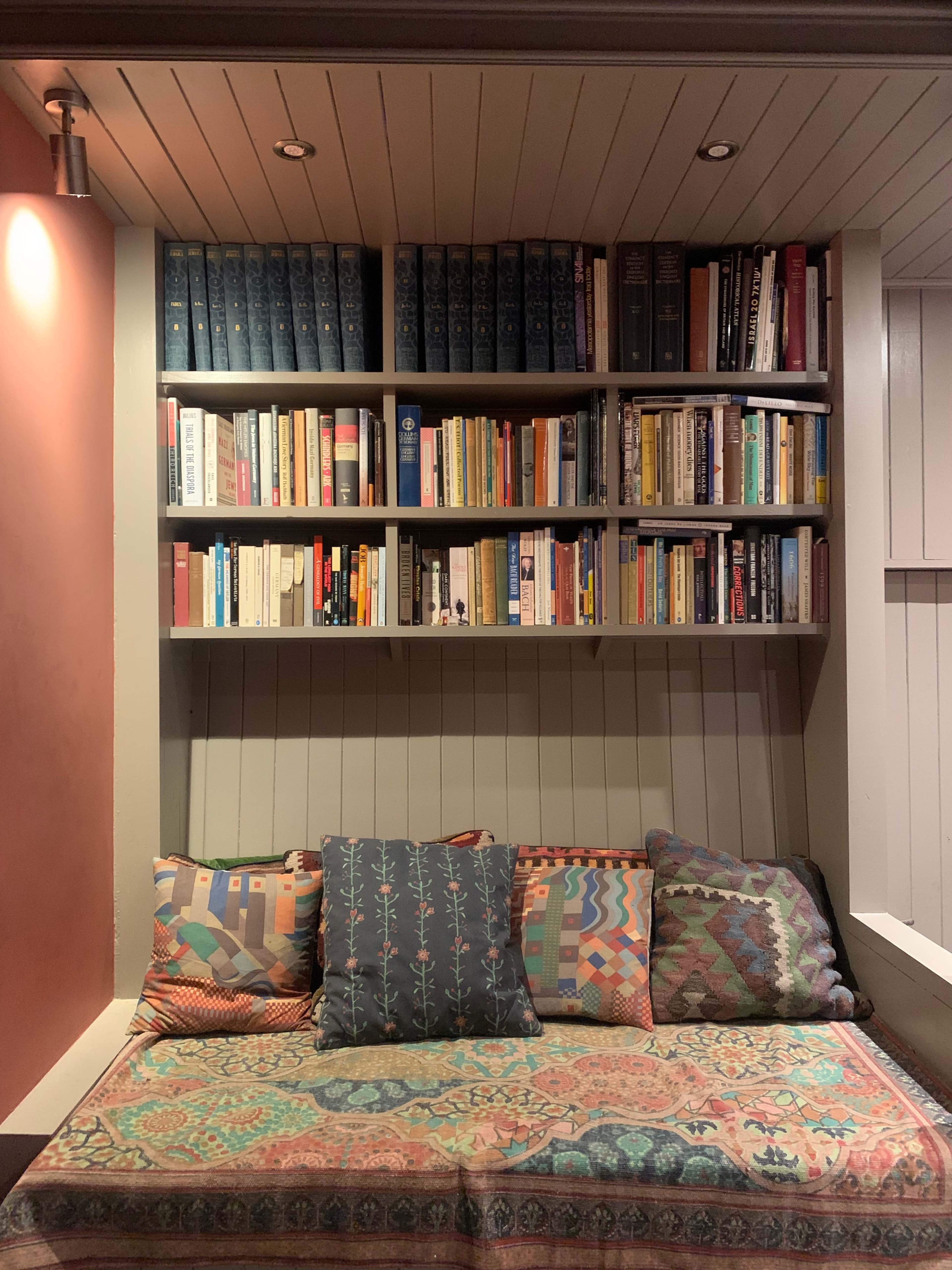The image shows a cozy nook with a bench covered in patterned pillows and a wall-mounted bookshelf filled with various books.