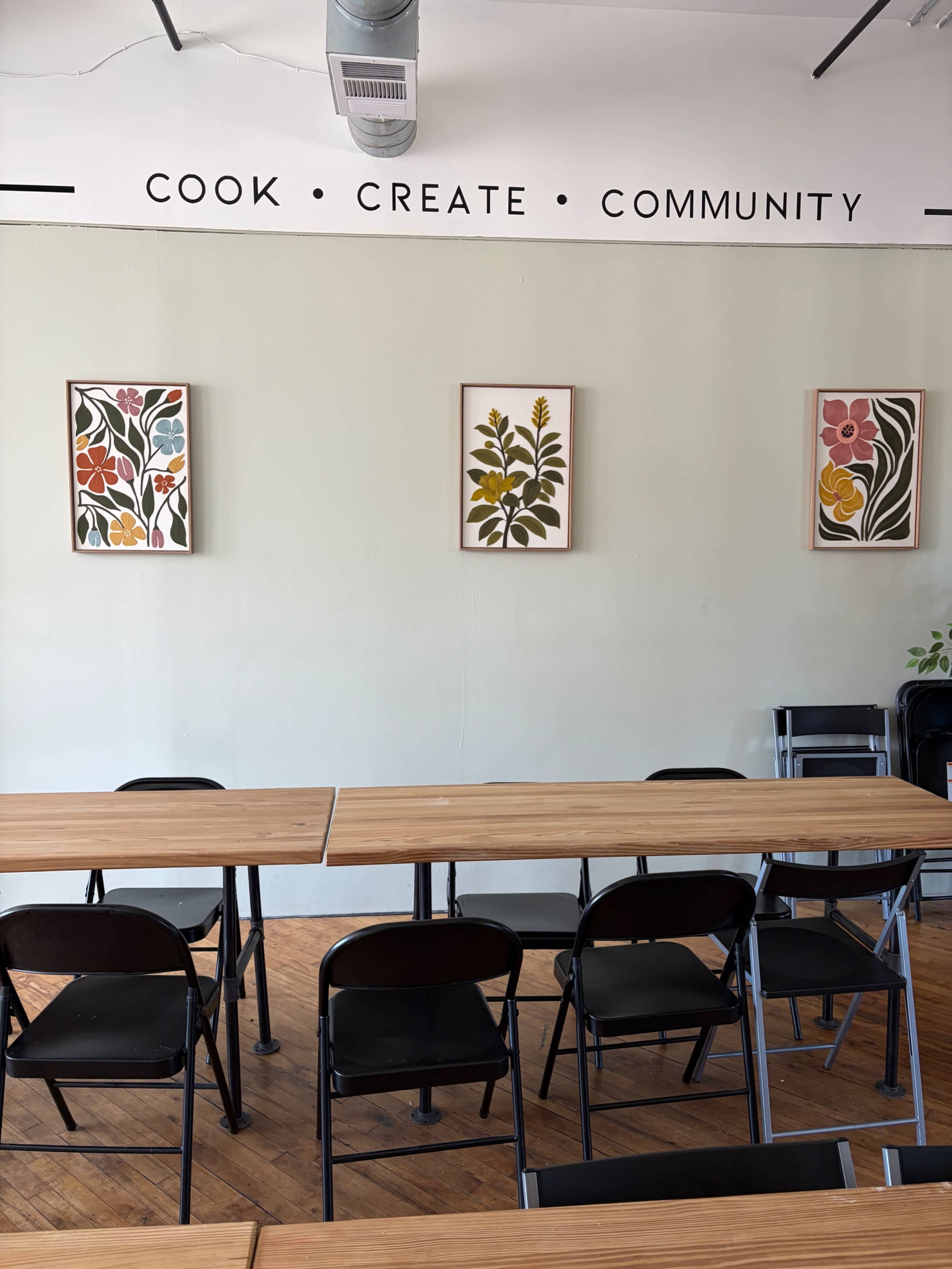 The image shows a room with three framed floral artworks on a light-colored wall, and several wooden tables surrounded by black chairs.