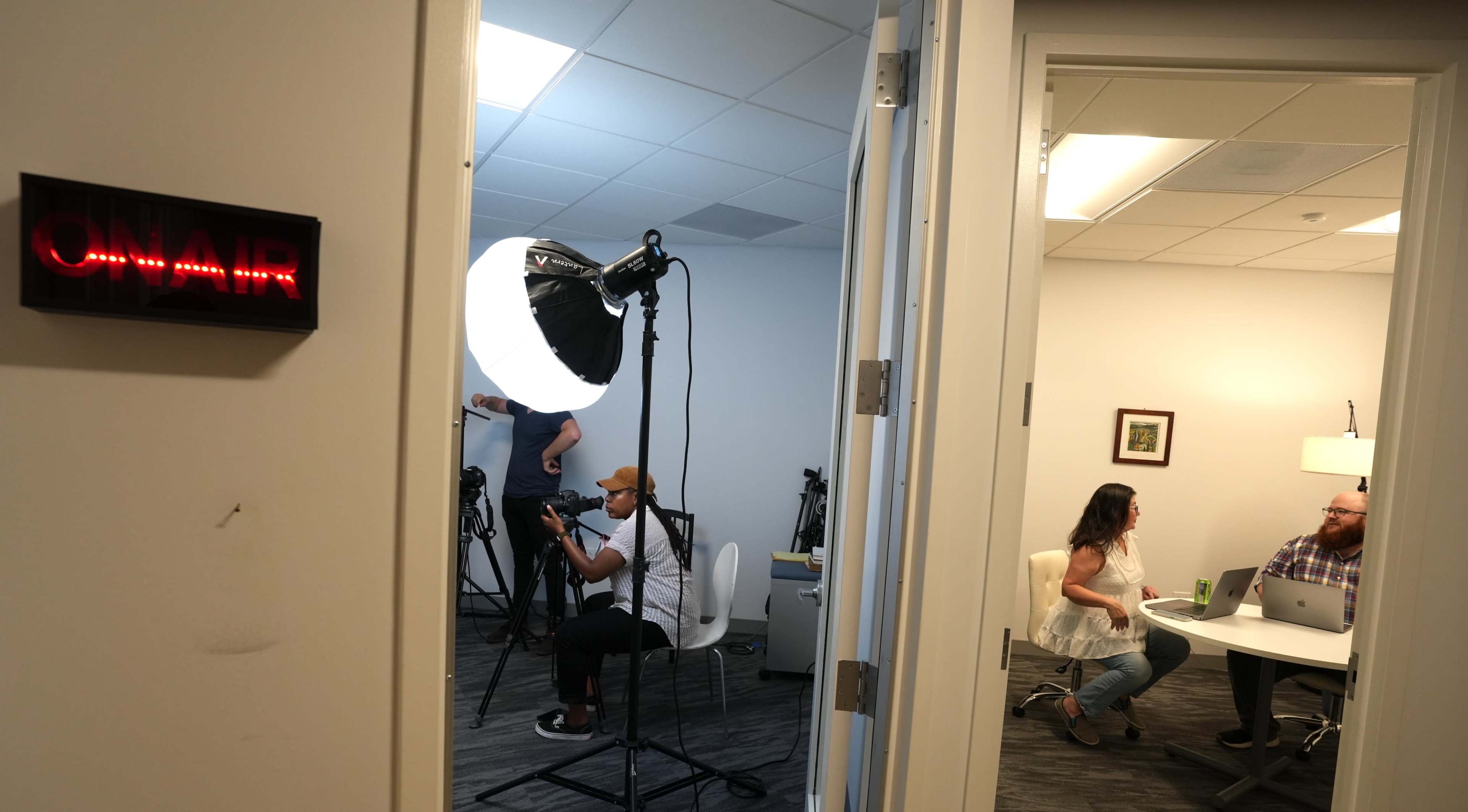 A person is sitting in a white chair preparing for an interview in a studio with a large light source, while another person works at a table in a nearby office.