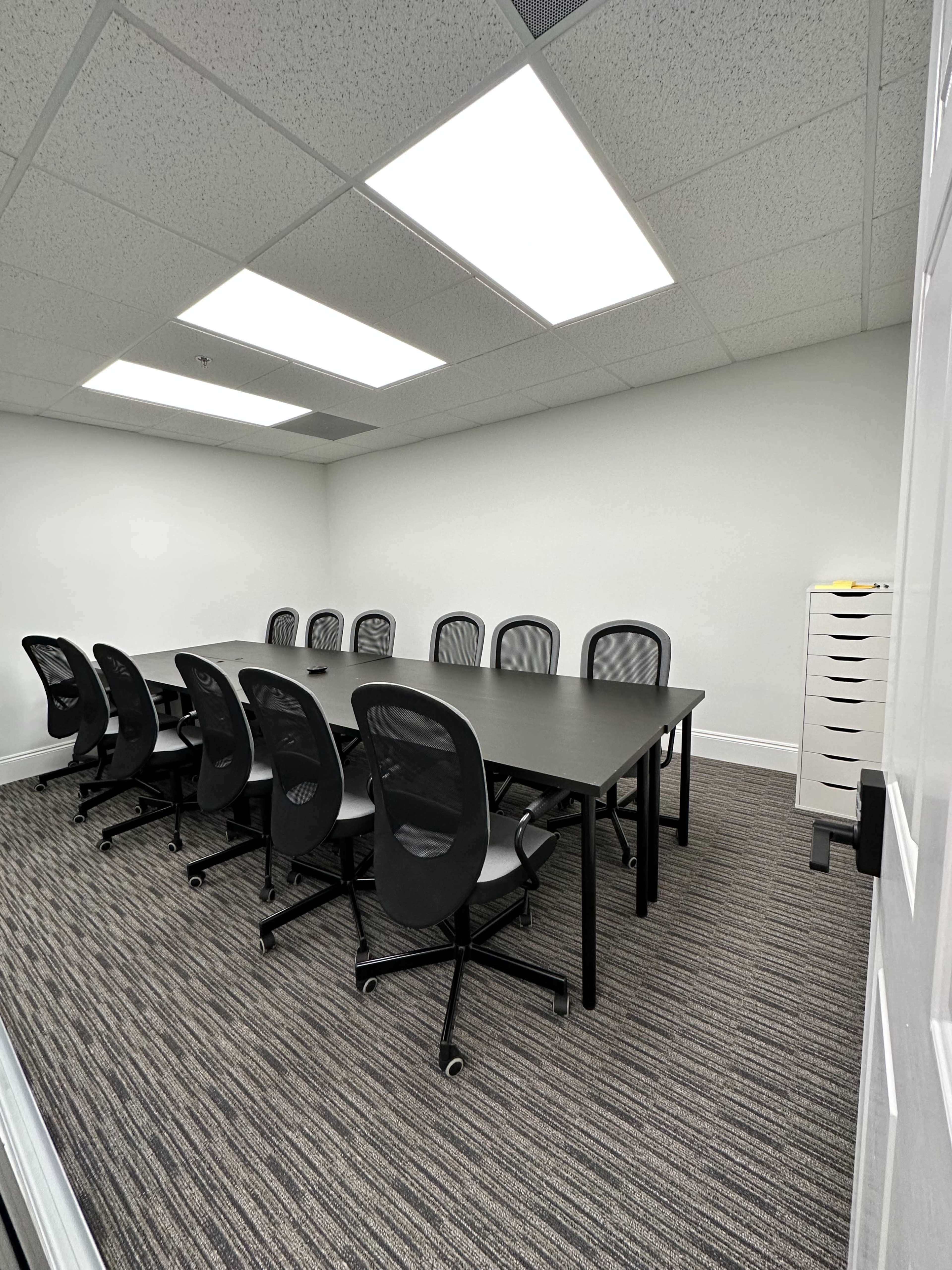 A conference room featuring a long table surrounded by eight black ergonomic chairs, with a storage cabinet in the corner and bright overhead lighting.