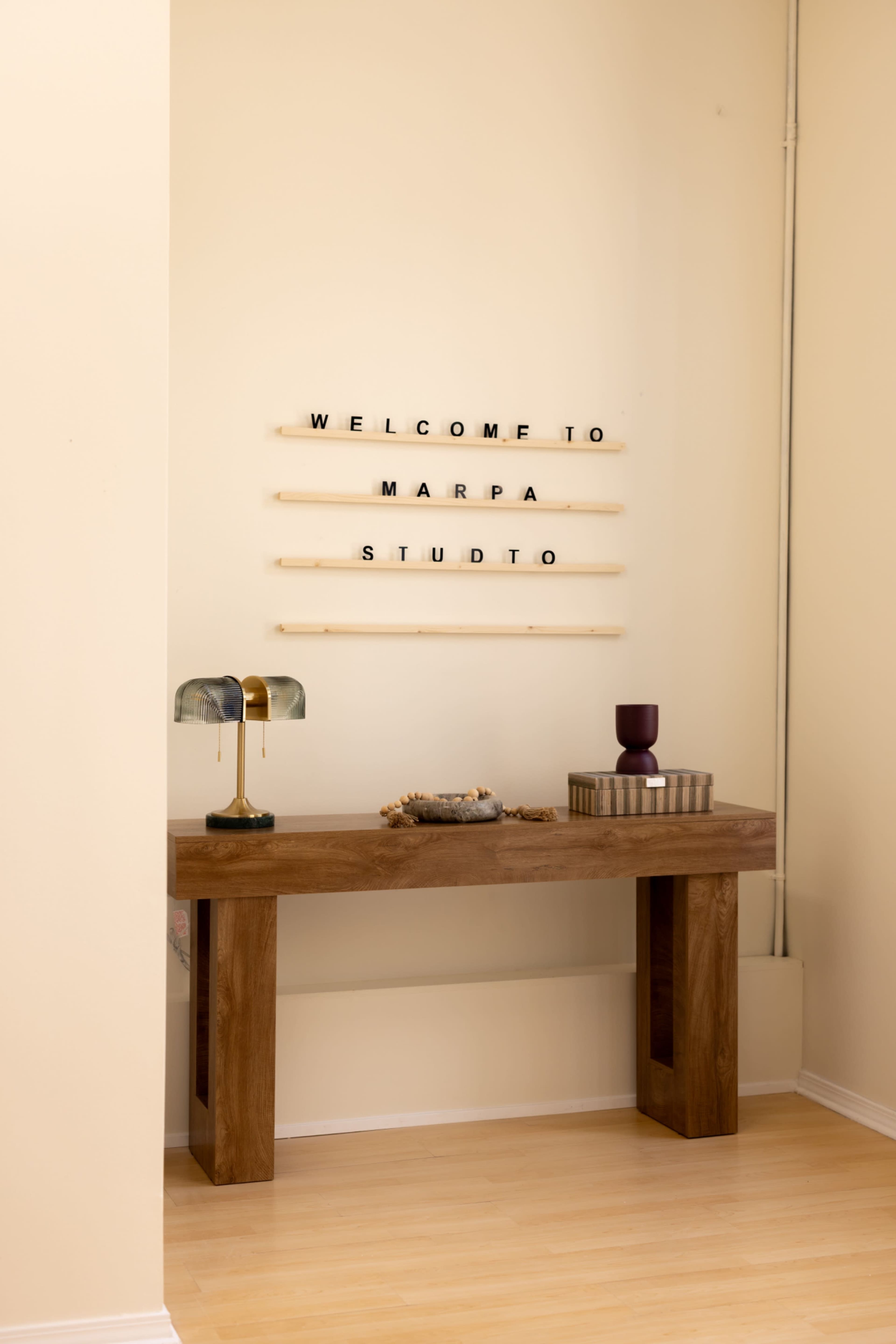 A wooden table with a lamp and decorative items is positioned against a wall displaying the text "WELCOME TO MARPA STUDIO" in black letters.