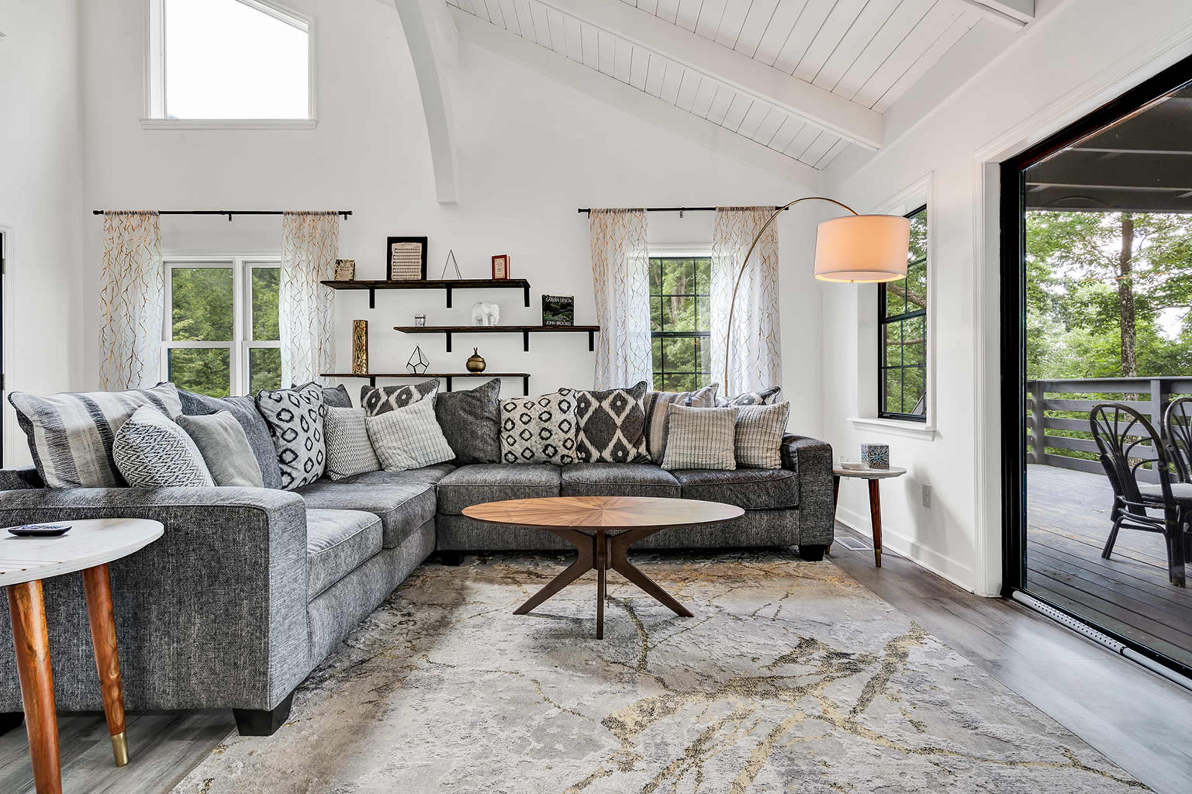 The image shows a modern living room with a sectional gray sofa, decorative pillows, a round wooden coffee table, and large windows opening to a view of trees outside.