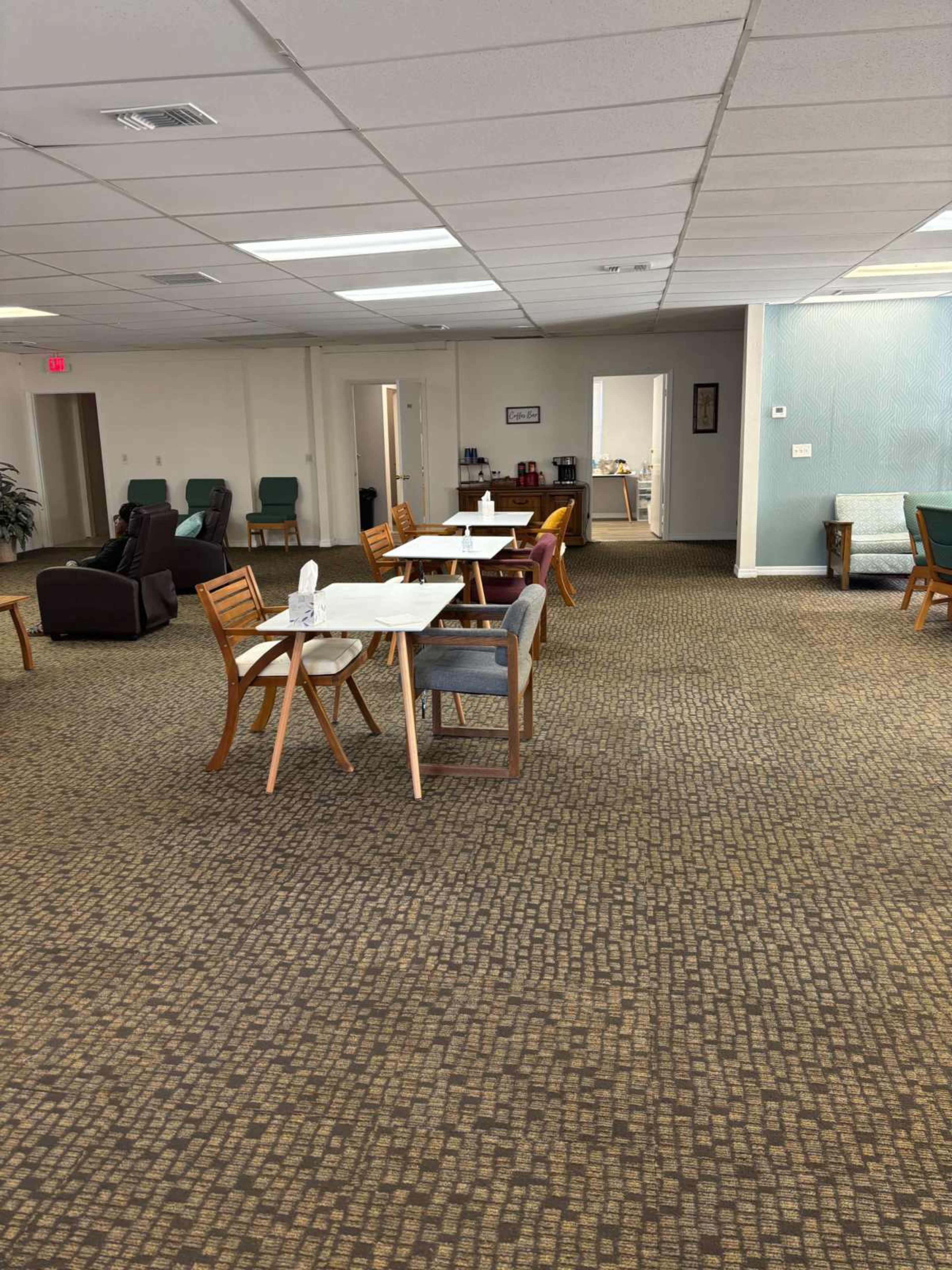 A spacious room with several tables and chairs arranged on a carpeted floor, leading to an open doorway in the background.