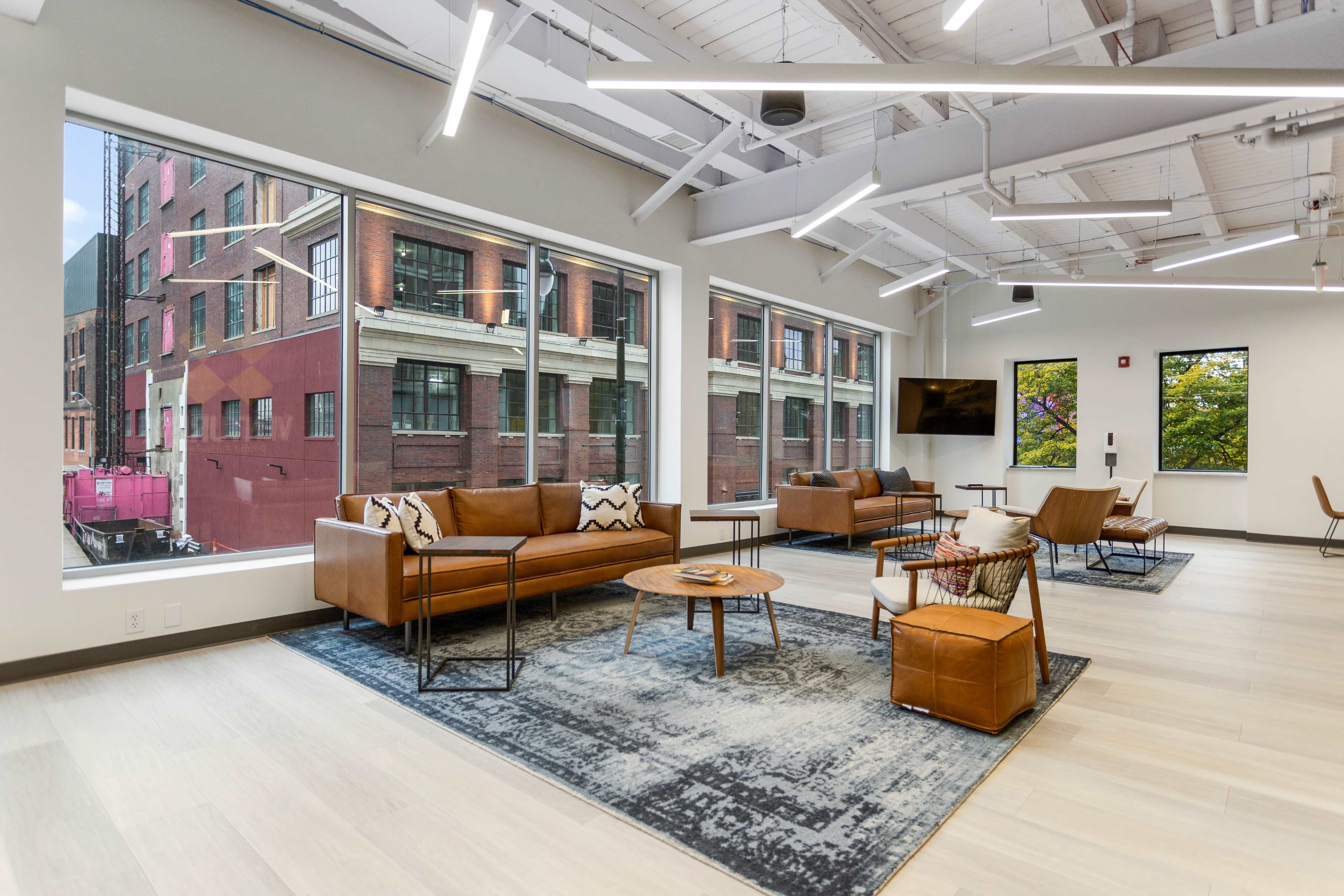 A modern lounge area with brown leather sofas and chairs, large windows displaying a city view, and a light-colored wooden floor.