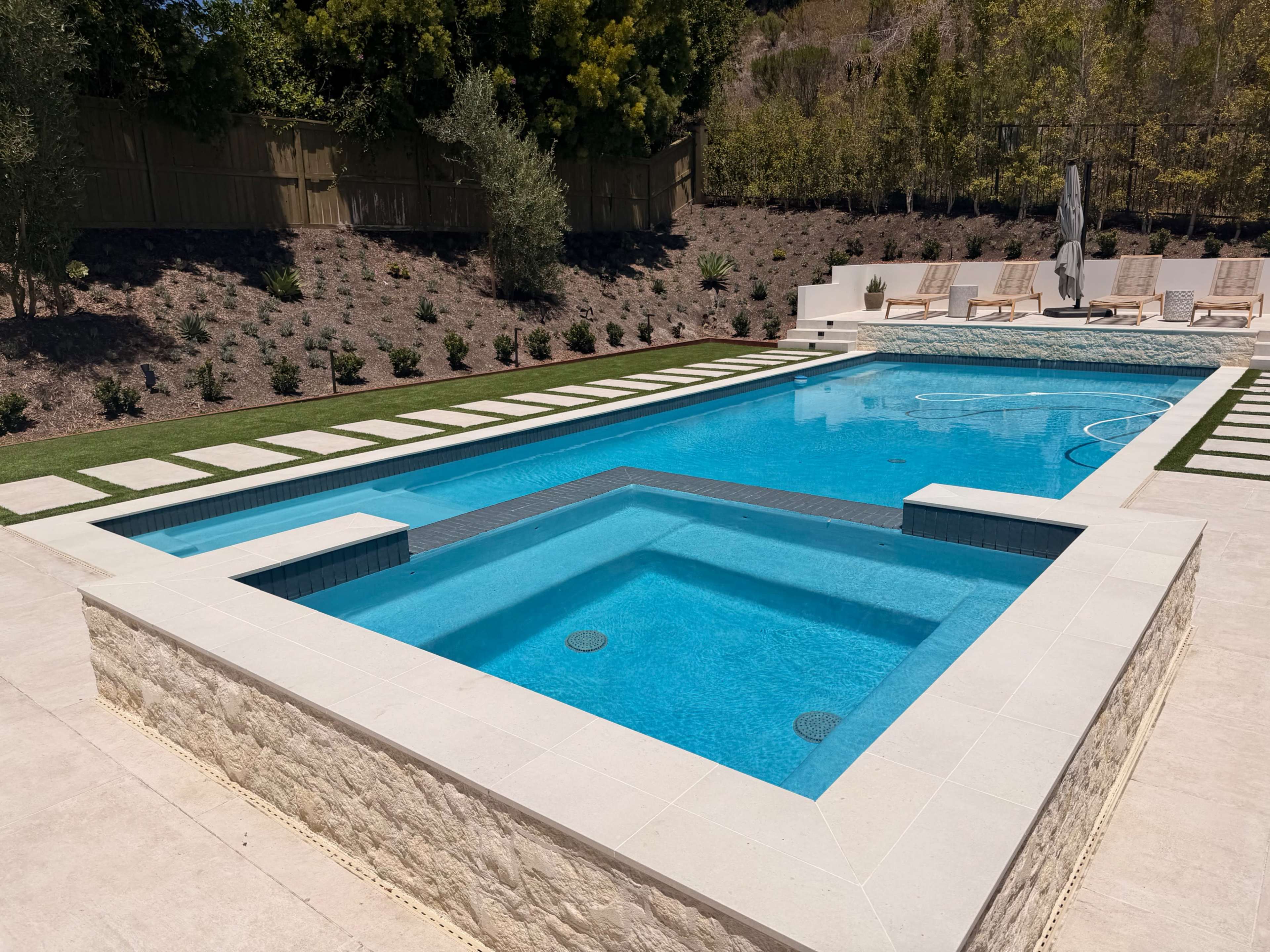 The image shows a rectangular swimming pool surrounded by landscaped greenery and a stone patio with lounge chairs.