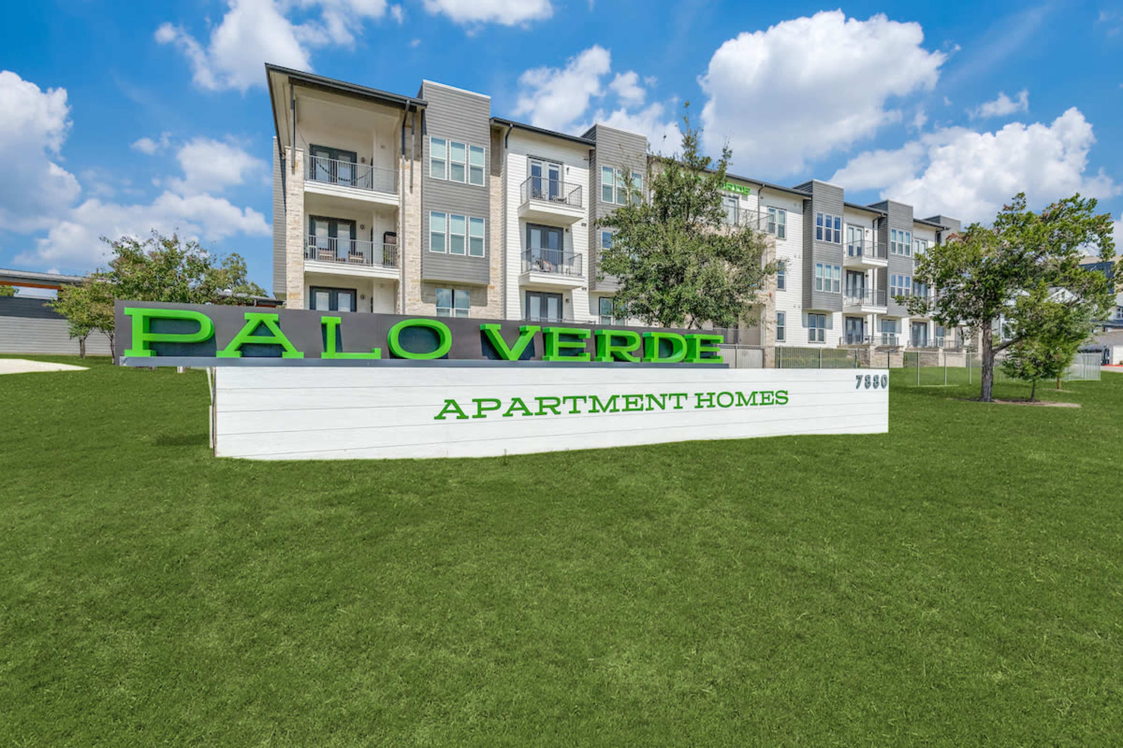 The image shows a large green sign reading "PALO VERDE APARTMENT HOMES" in front of a three-story apartment building surrounded by grass and trees.