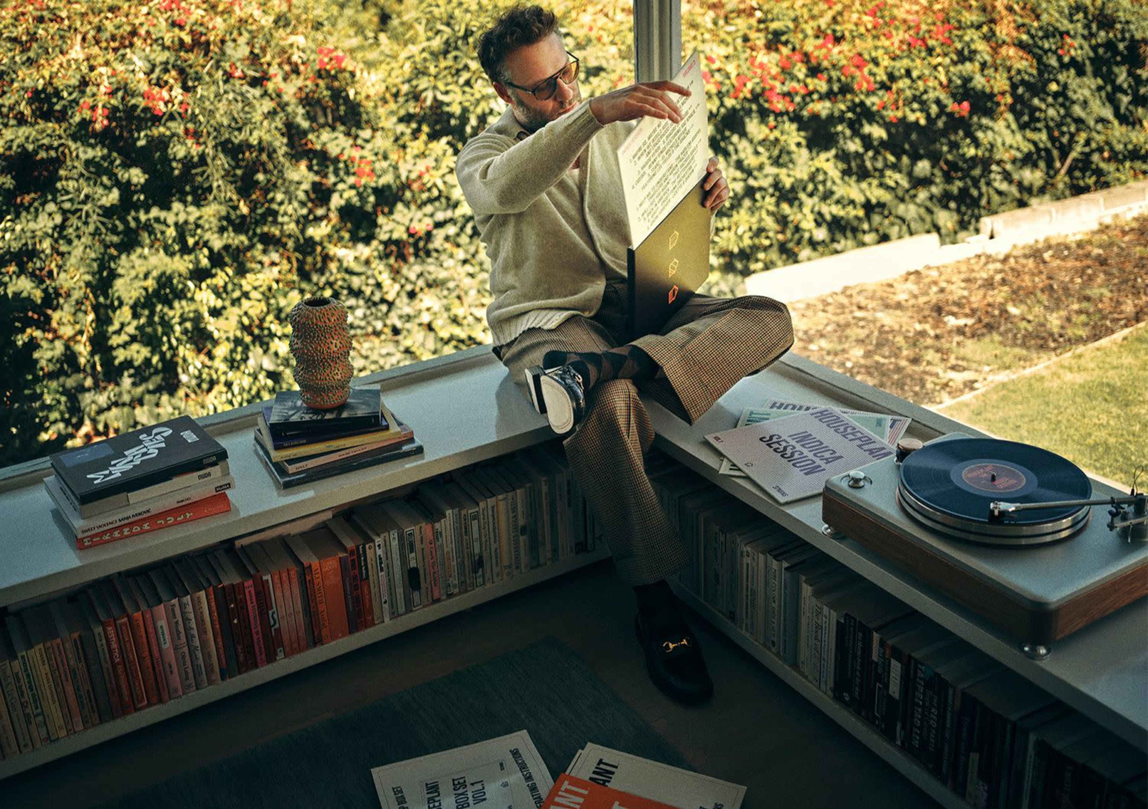 A man sits cross-legged on a shelf, examining a vinyl record while surrounded by books and a turntable in a sunlit room.