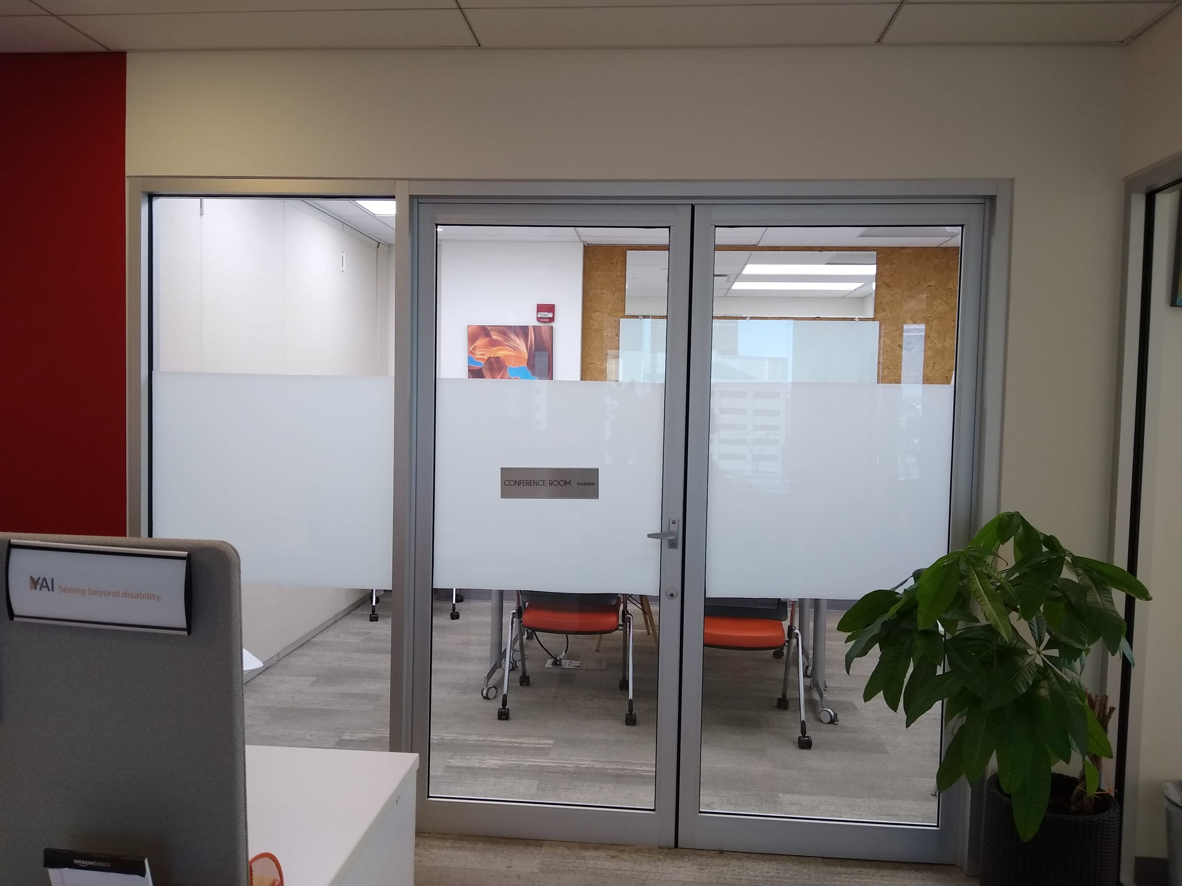 The image shows a glass-walled conference room with orange chairs and a plant in the foreground.