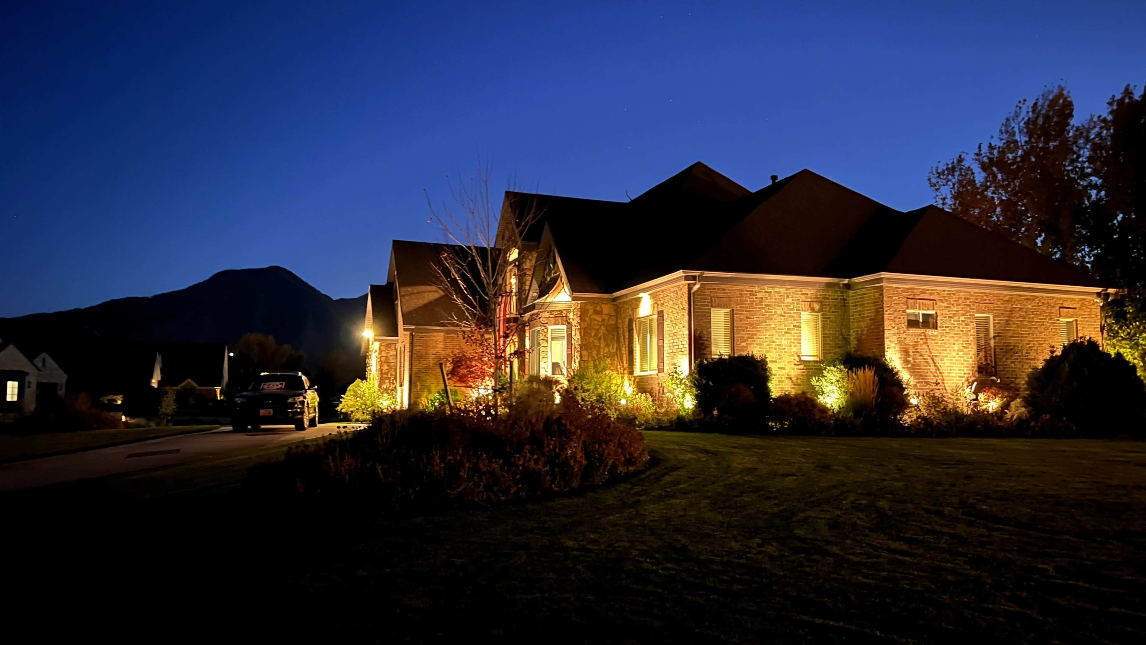 A well-lit brick house is surrounded by neatly landscaped gardens and set against a backdrop of a darkening sky and distant mountains.