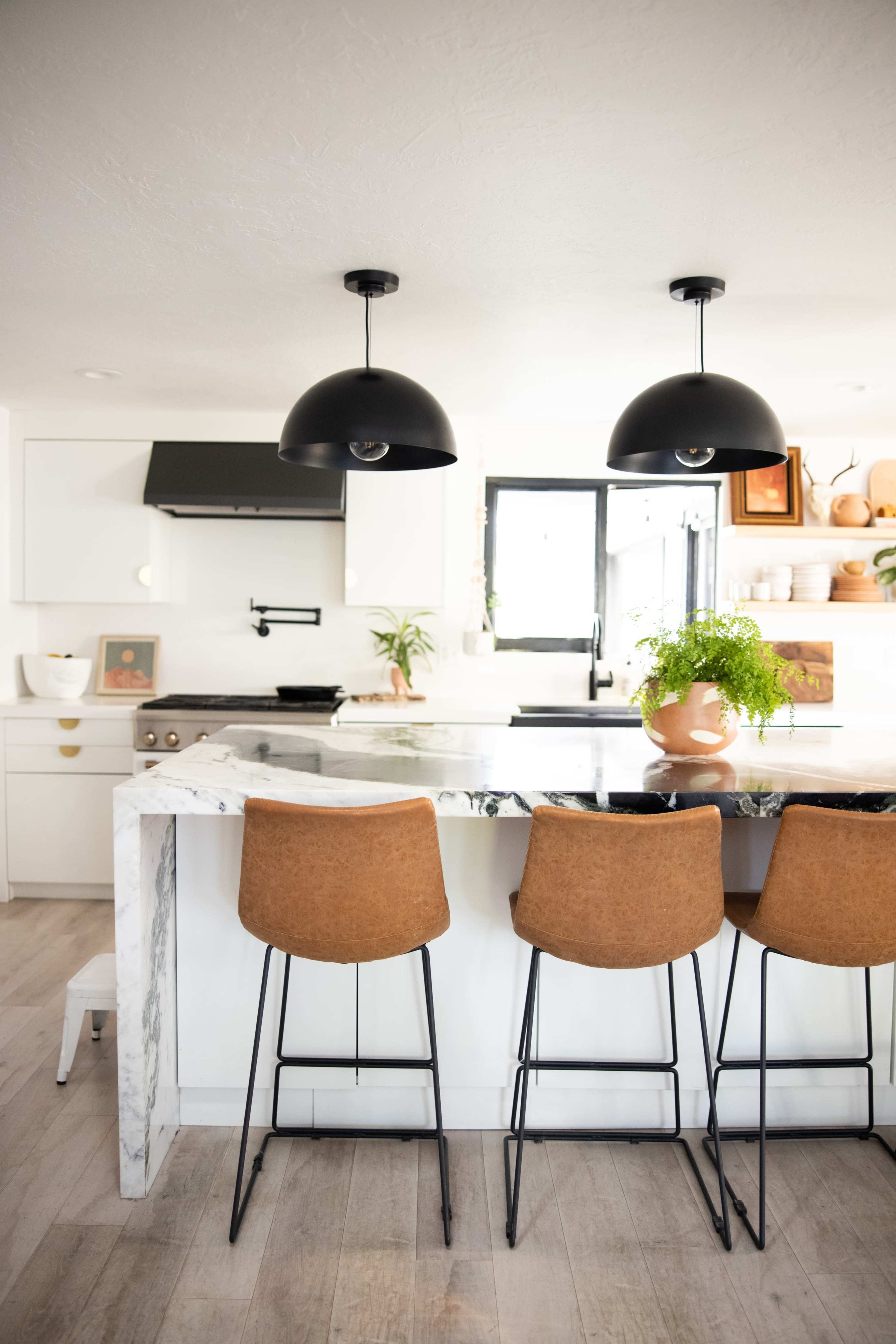 A modern kitchen features a marble-topped island with three brown bar stools and two black pendant lights hanging above.