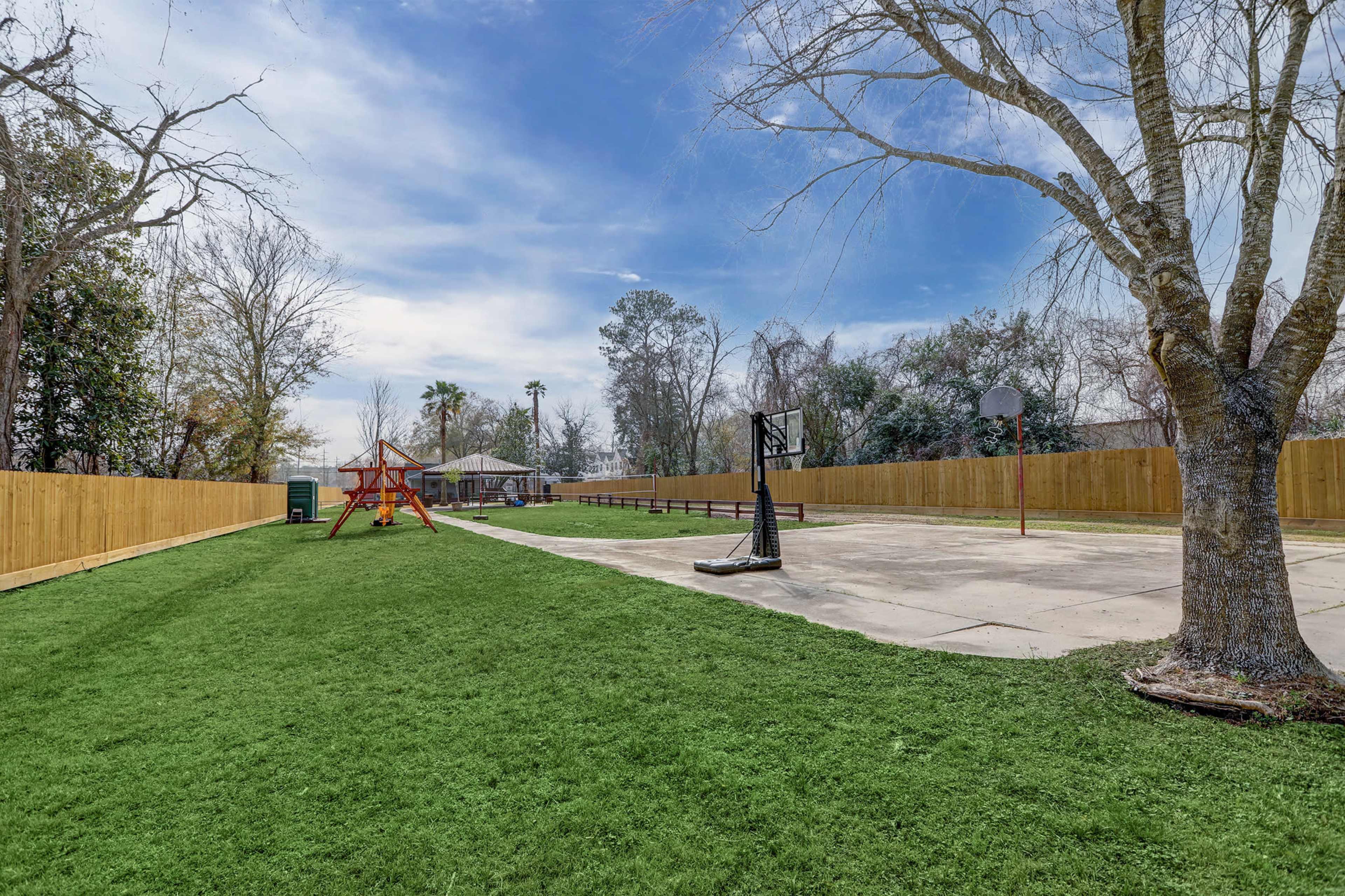 A fenced outdoor area with a grassy lawn, a playground structure on one side, and a basketball court on the other.
