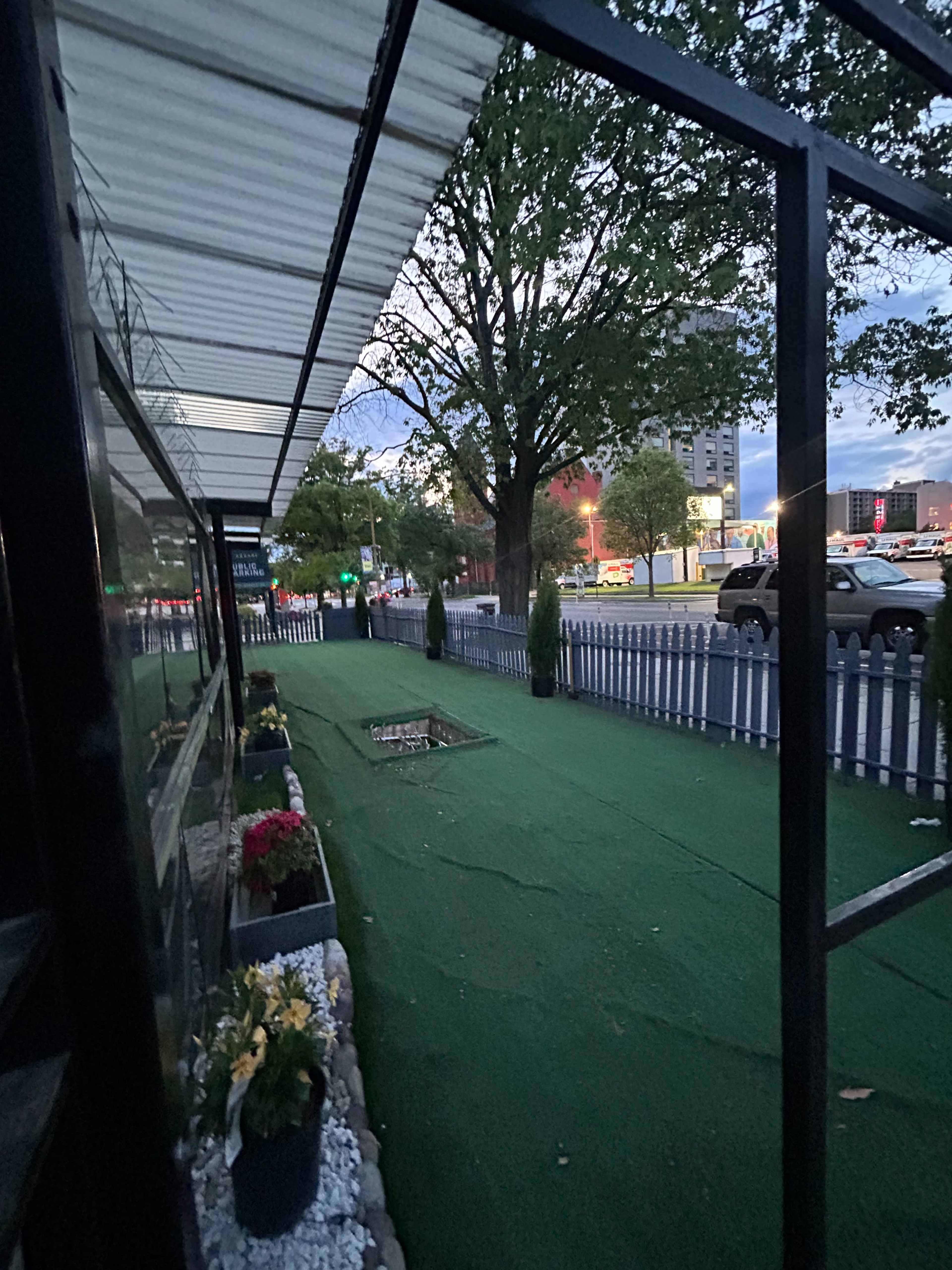 The image shows a walkway lined with planters and a picket fence, leading to a road with traffic in the background during dusk.