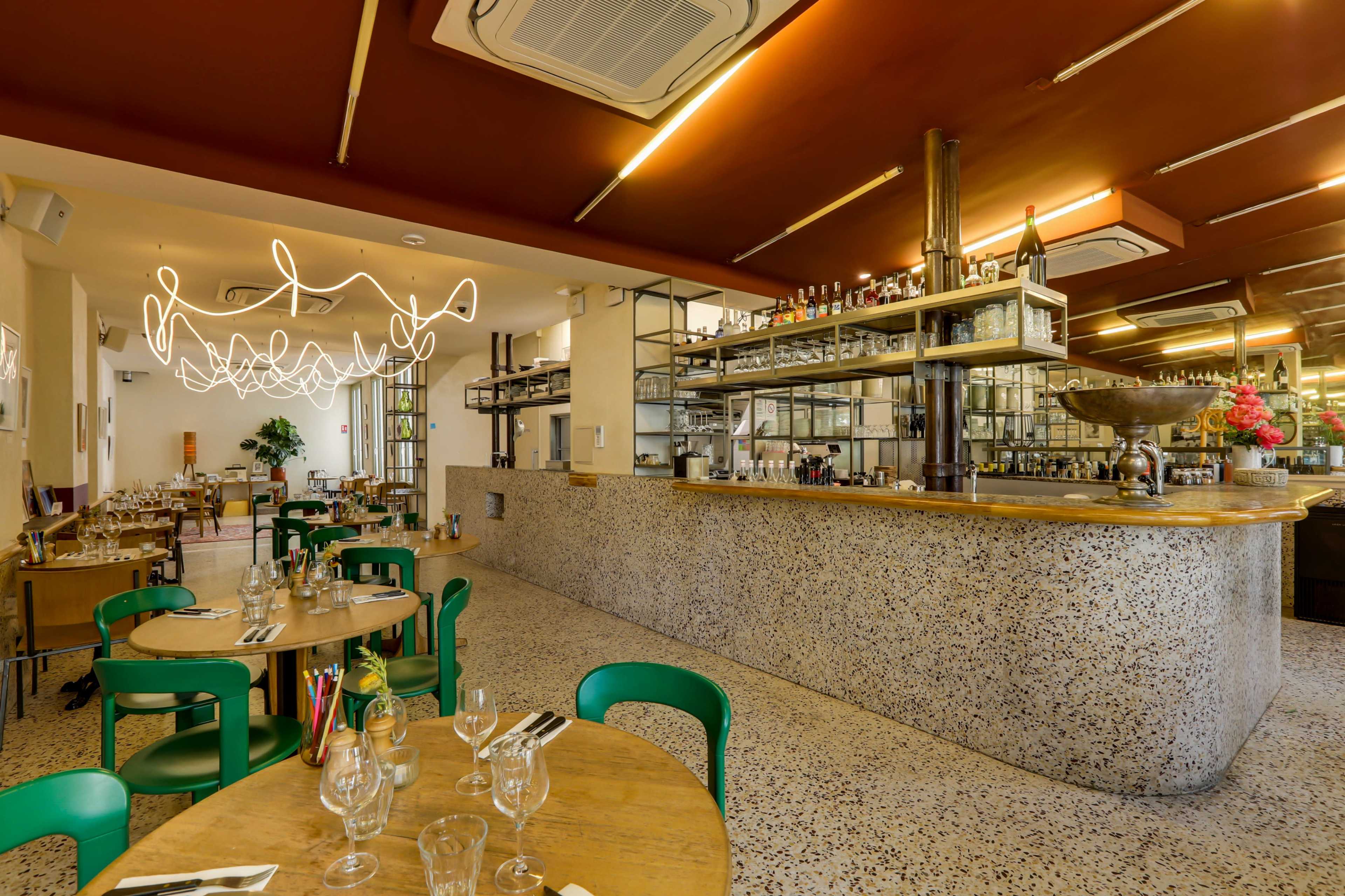 The image shows a modern restaurant interior featuring wooden tables with green chairs, a bar area with glass shelves displaying various bottles, and stylish overhead lighting.