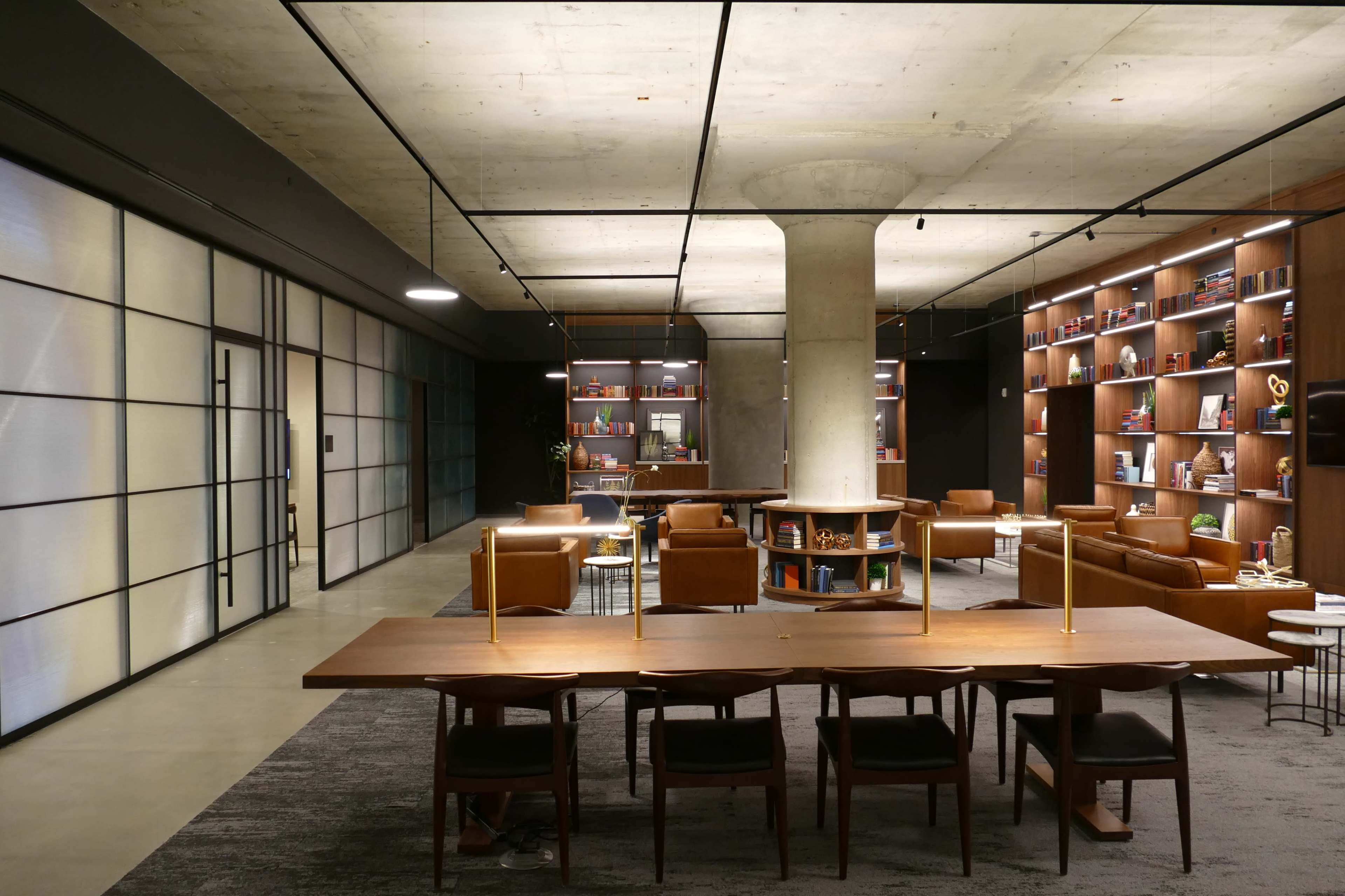A modern office space featuring a large wooden table with chairs, leather seating areas, and bookshelves along the walls.